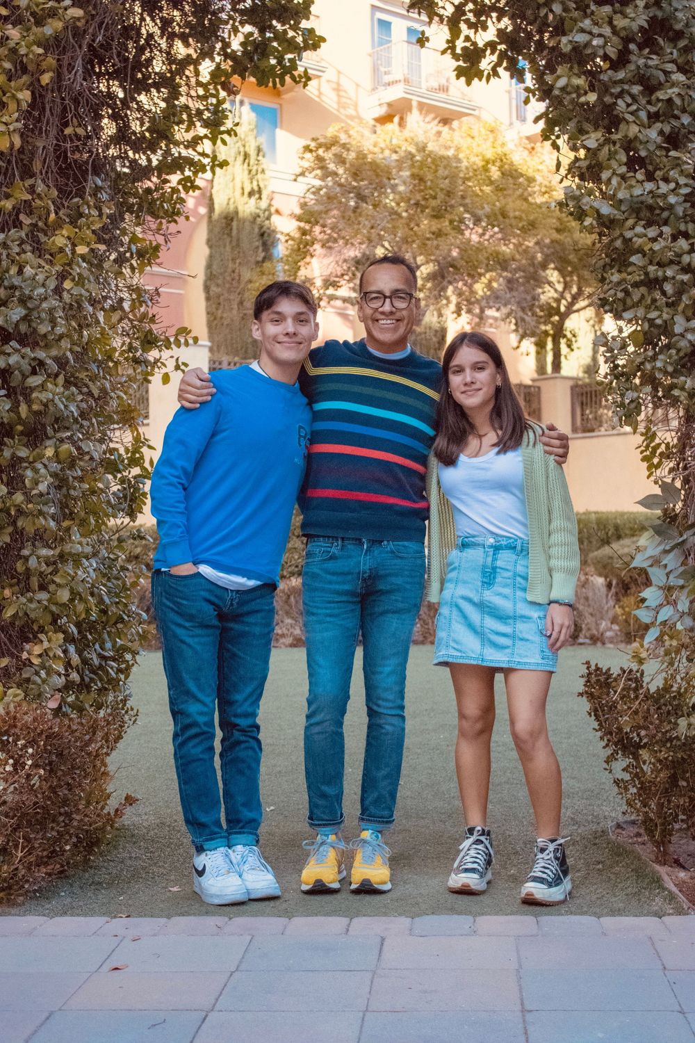A father in a striped sweater stands with his two teenagers under a leafy archway outdoors.
