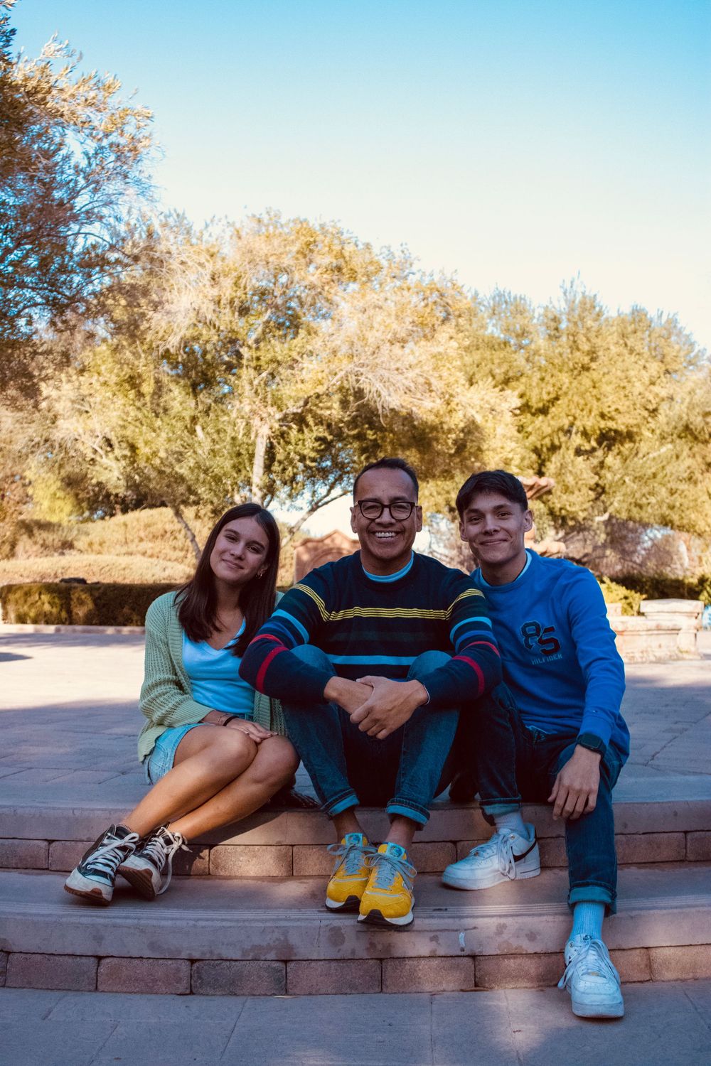 A father and his two teenage children sit together smiling on stone steps in a sunny park.