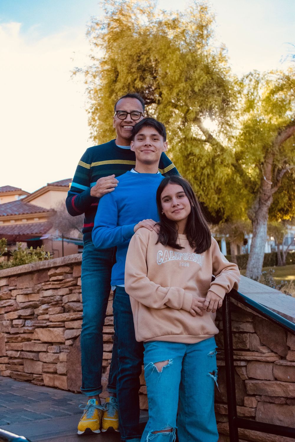 Father posing proudly with teenage son and daughter on stone wall terrace at sunset outdoors.