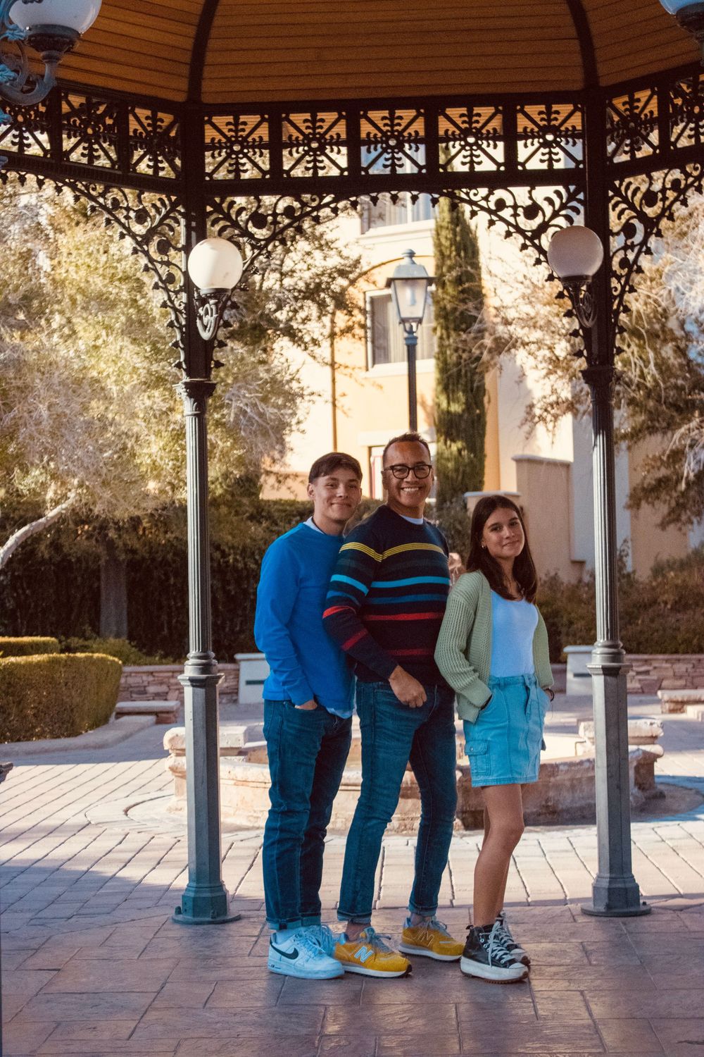 A father and two teenagers smile together under an ornate gazebo in a sunlit outdoor plaza.
