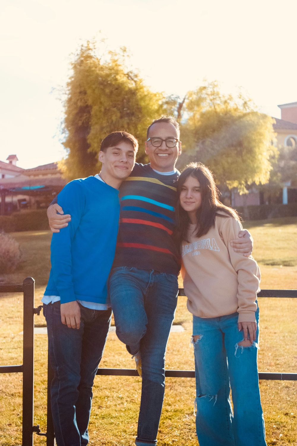 Three people embracing and smiling outdoors near a fence with golden autumn trees behind them.