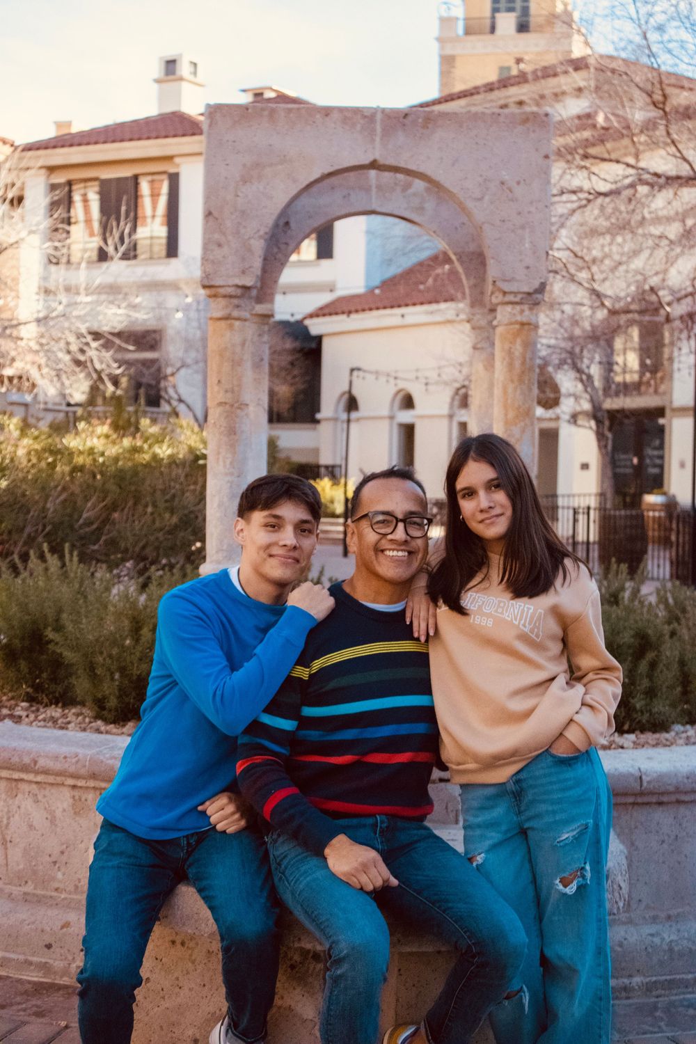 Three people posing together in front of a stone arch landmark at sunset.