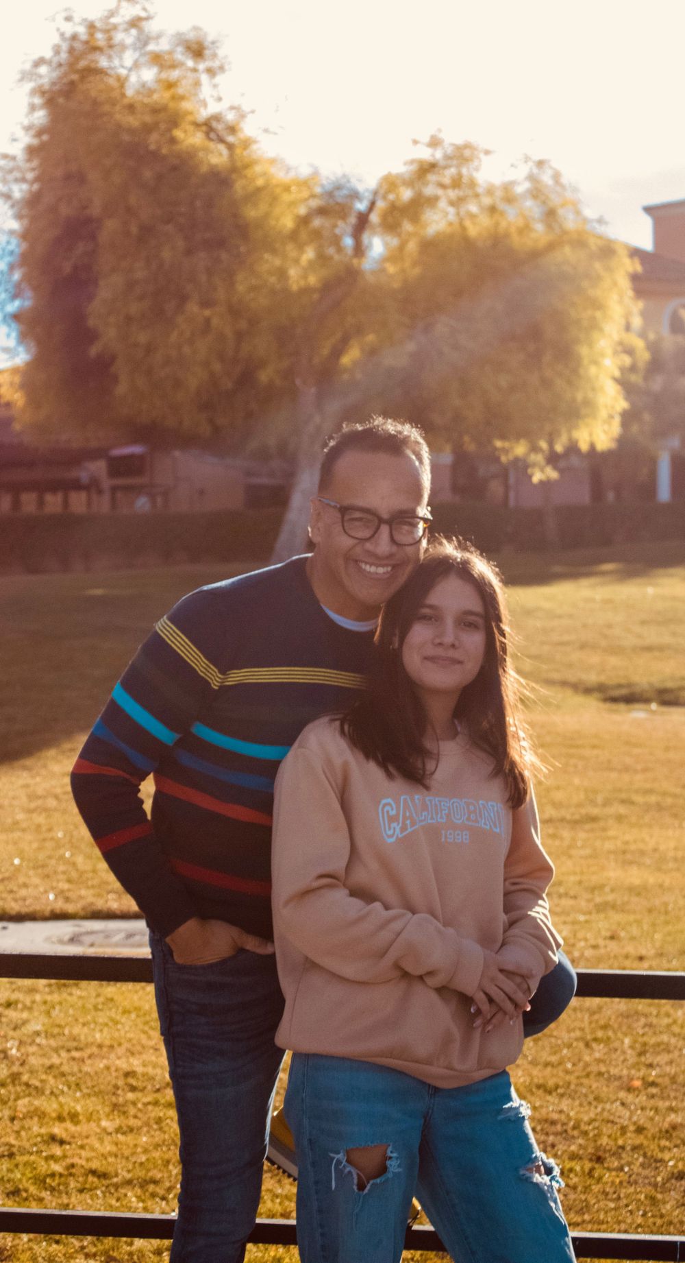Father and teenage daughter hugging outdoors in a park during golden hour sunset.
