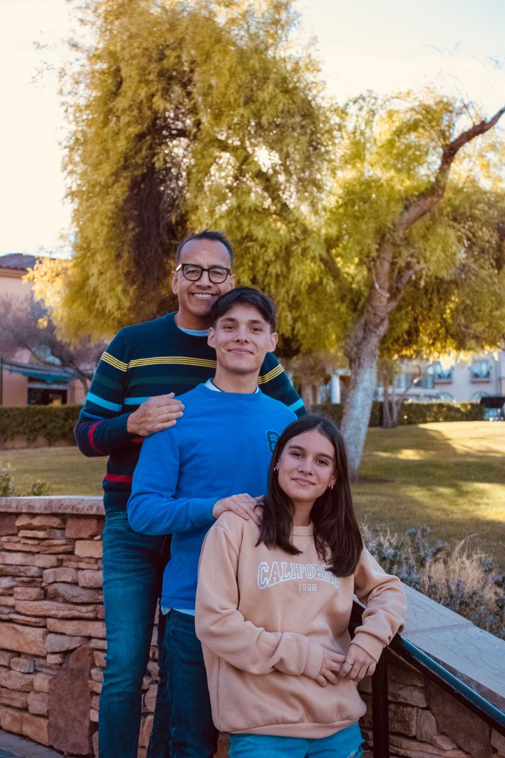 Father posing with teenage son and daughter outdoors in a park at golden hour.
