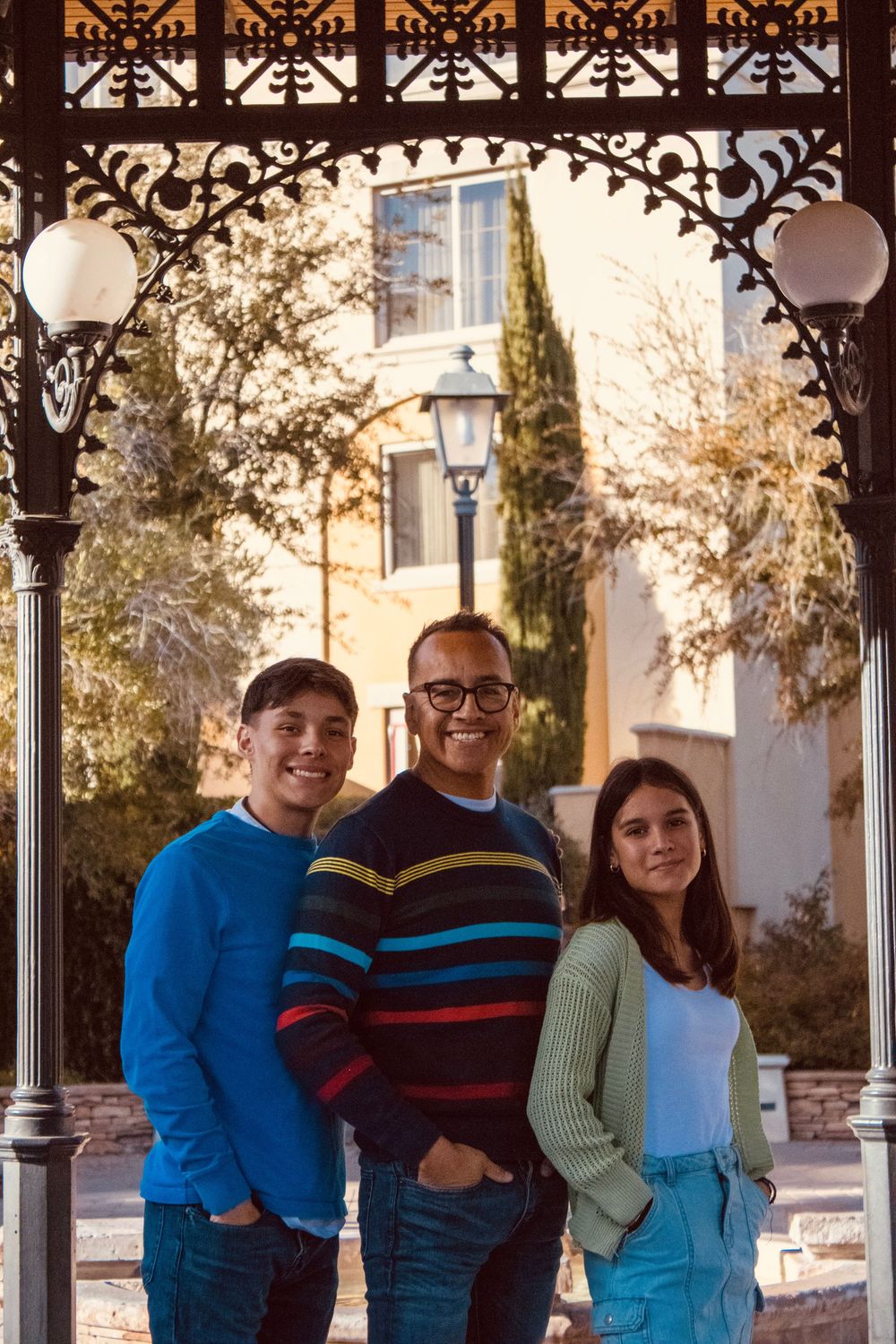 A man and two teenagers pose together under an ornate iron archway with warm golden-hour lighting behind them.