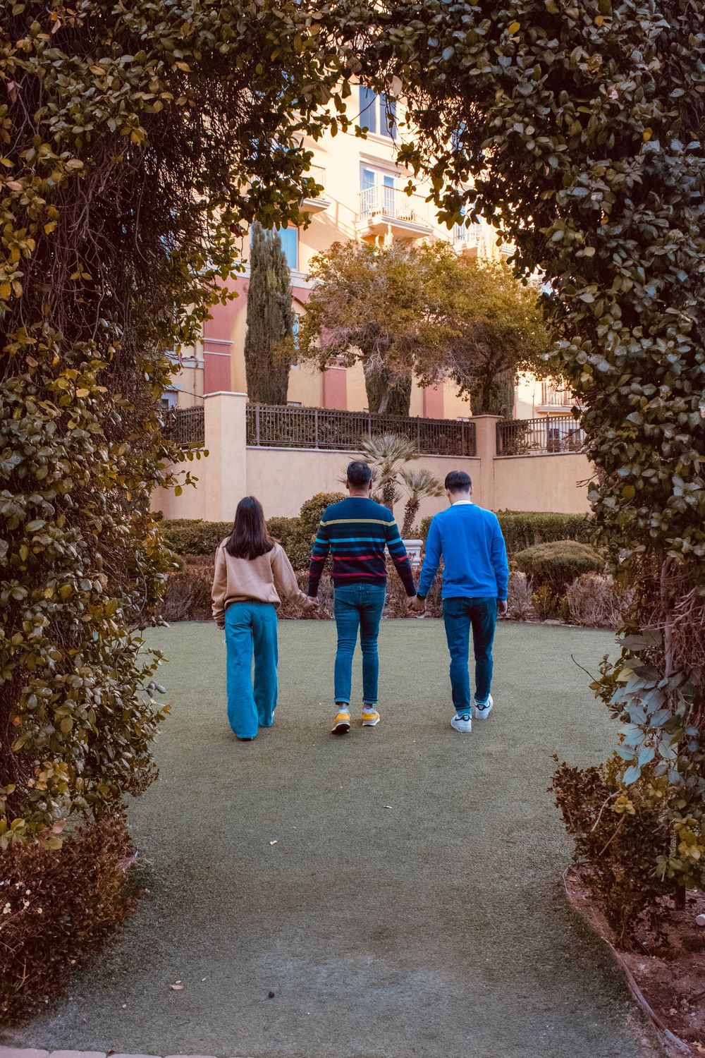 Three people walk hand-in-hand through a leafy garden archway toward Mediterranean-style buildings at sunset.
