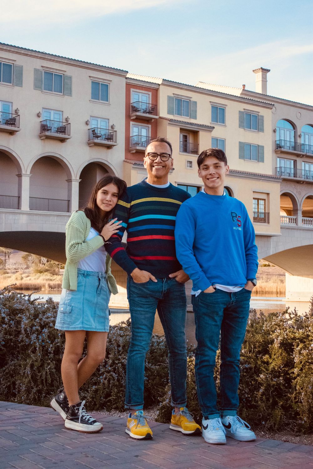 A father and his two teenagers smile together near a waterway with arched bridge and blue sky behind them.