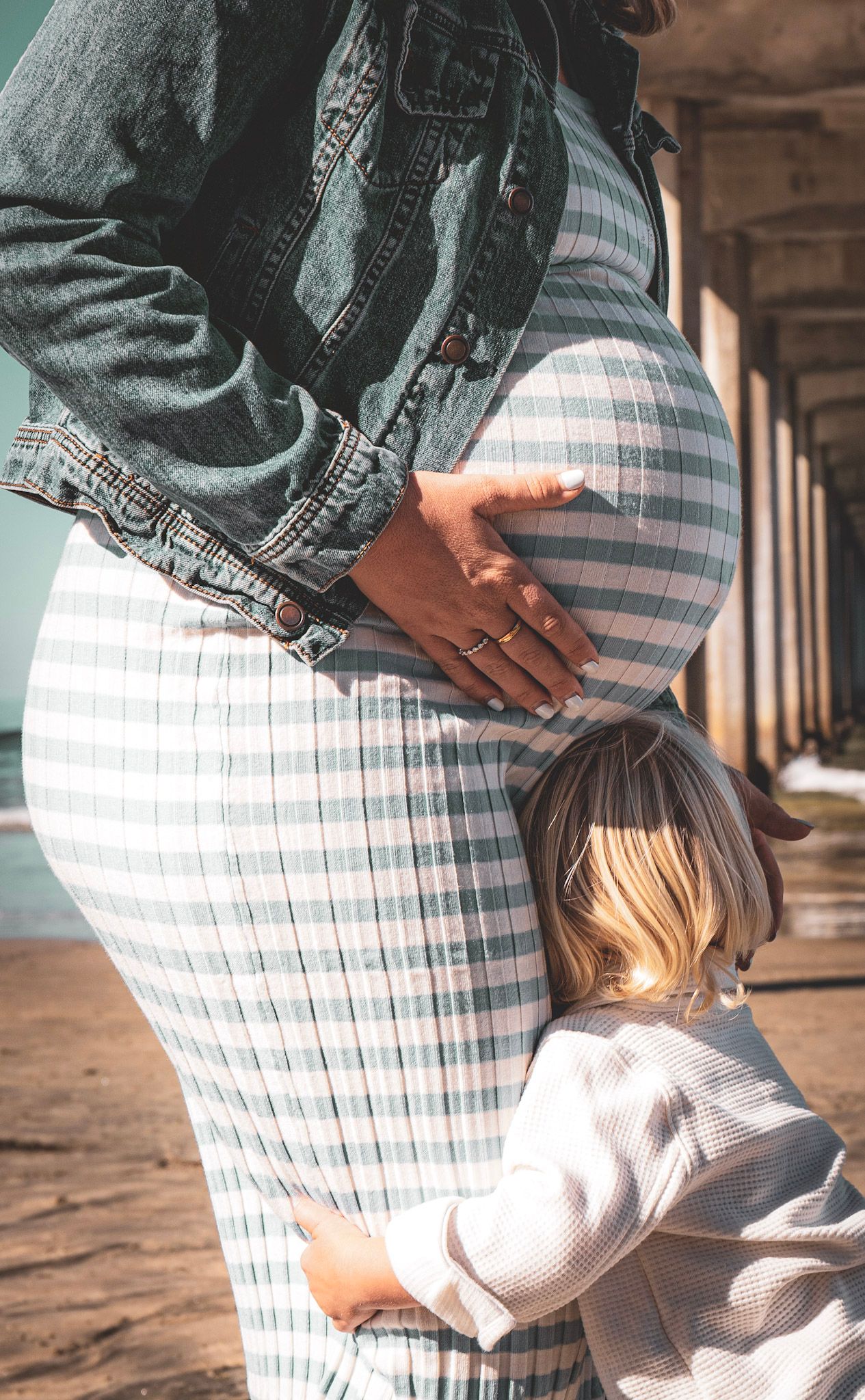 Pregnant woman in striped dress with small child hugging her belly in a hallway.