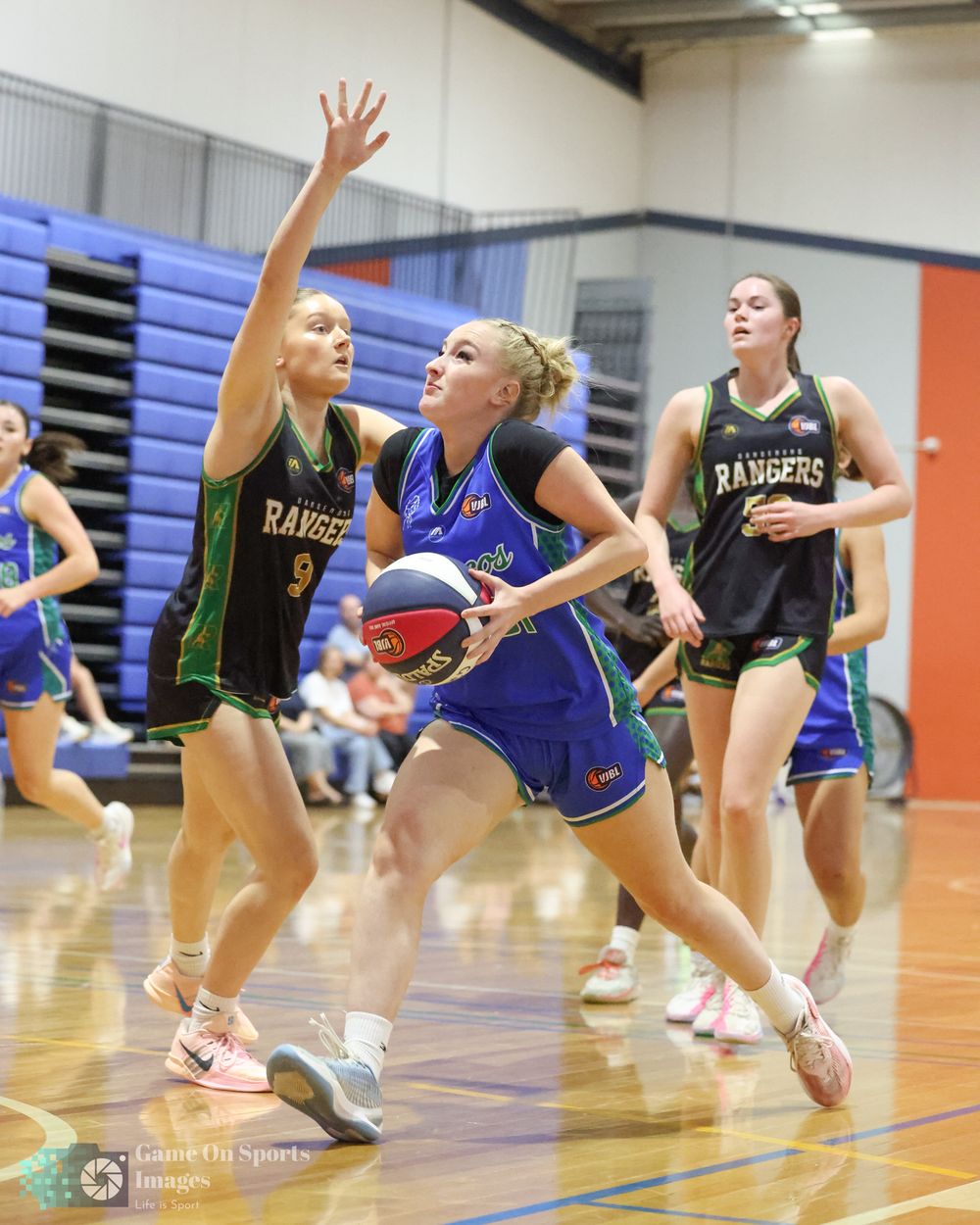 Female basketball player in blue drives to the basket against defenders in black Rangers uniforms during an indoor game.