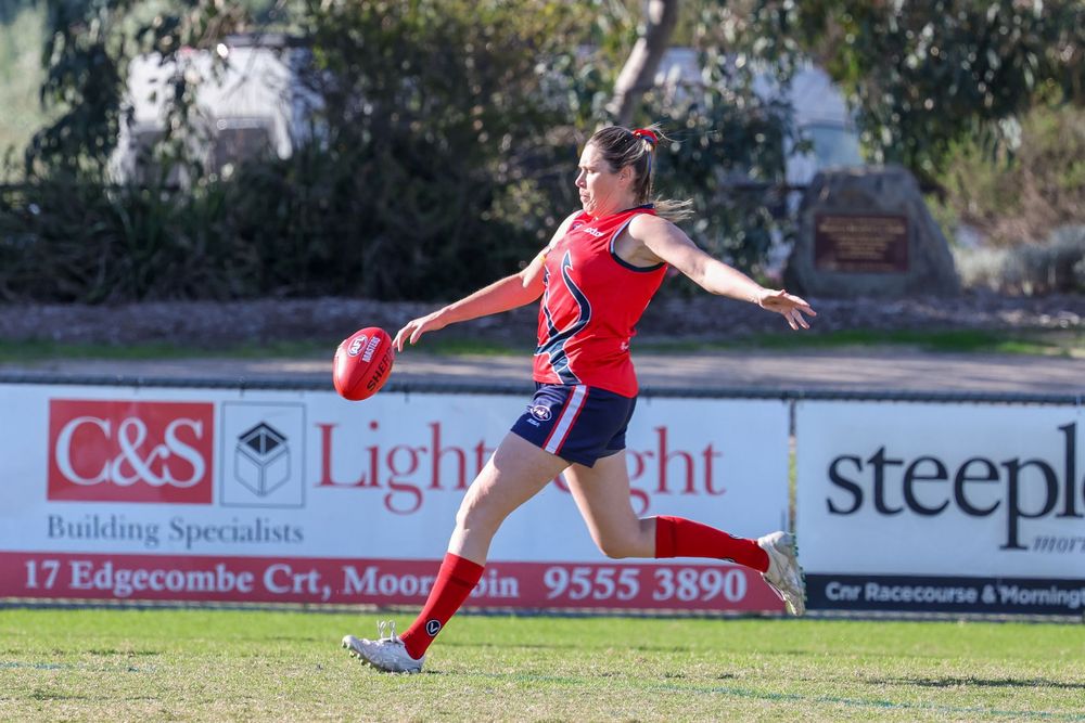 Female Australian rules footballer in red and blue uniform kicking ball during match with sponsor signage in background.