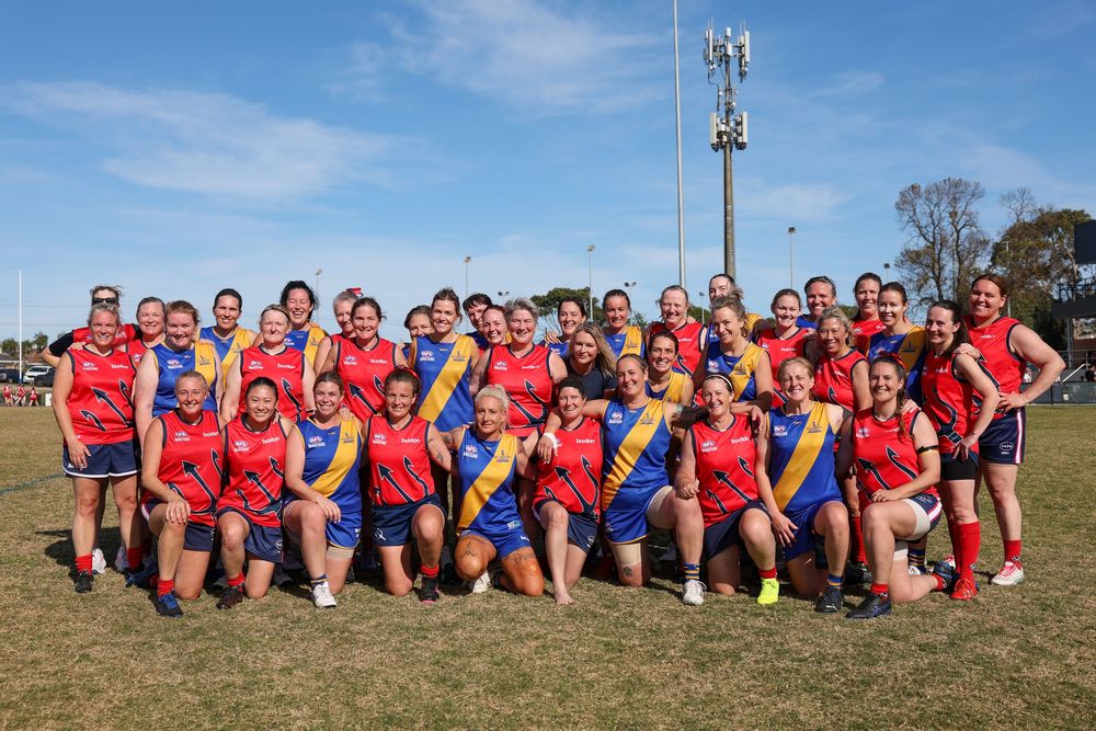 Women's Australian rules football teams posing together on a sunny oval after a match.