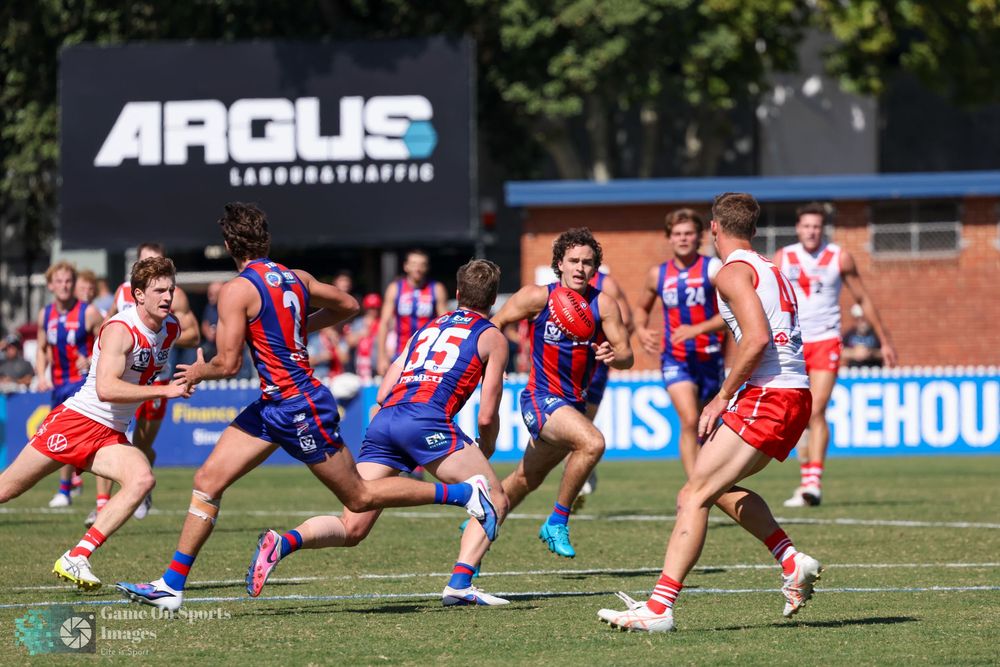 AFL players in red, white, and blue uniforms compete on a sunny oval with an Argus Laboratory Traffic sign in the background.