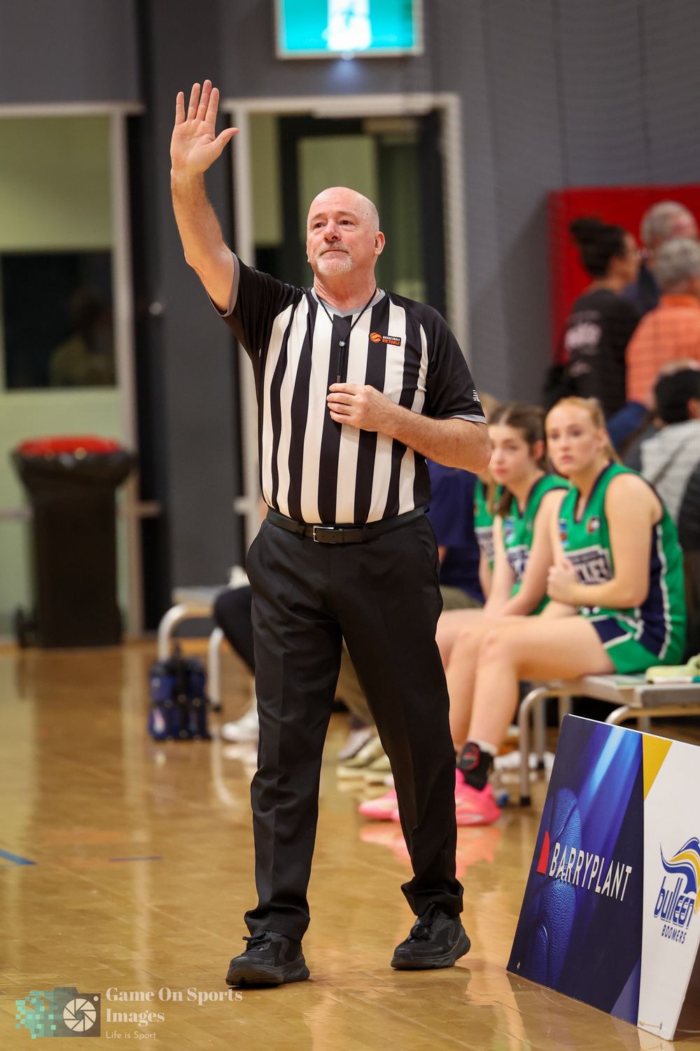 A bald basketball referee in black and white stripes raises one hand to signal a call during an indoor game.