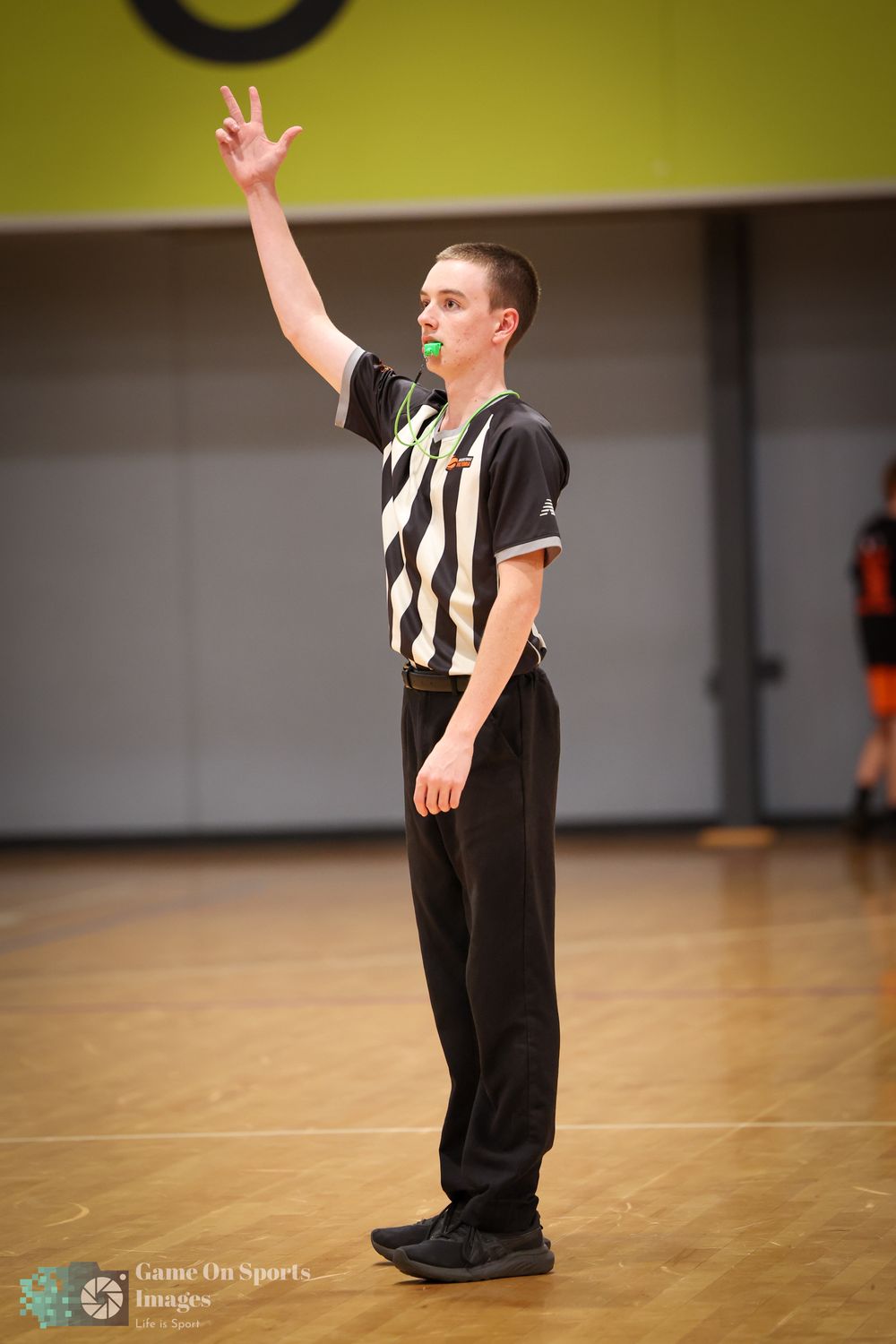 Young basketball referee in black and white striped shirt raising hand to signal a call on indoor court.