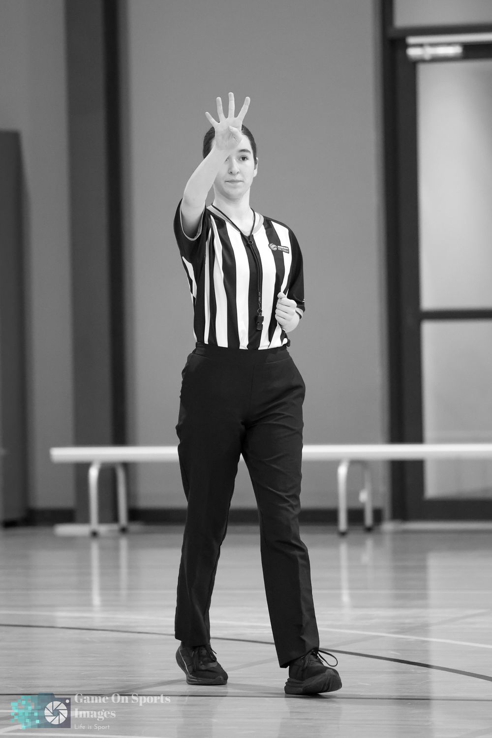 Female referee in striped shirt signals with raised hand on indoor basketball court.
