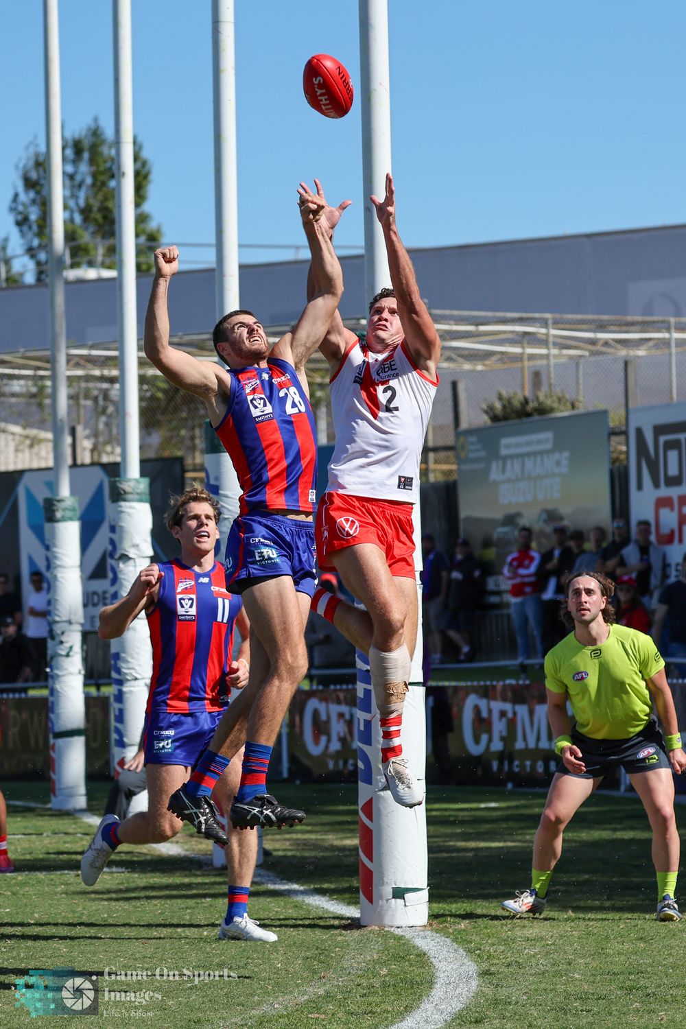 Two Australian rules footballers leap to contest a mark near the goal post during a competitive VFL match on a sunny day.