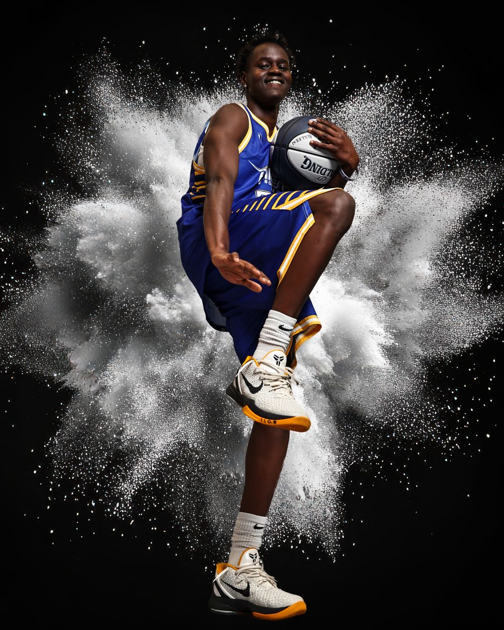 Basketball player in blue uniform poses dynamically with ball amid white powder explosion on black background.