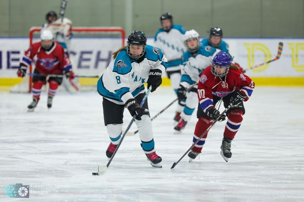 Two ice hockey players compete for the puck during an indoor rink game, wearing teal and red uniforms.