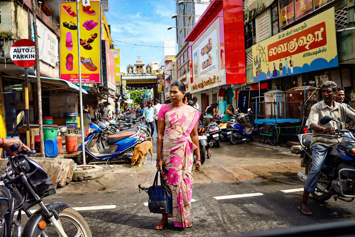 Woman in pink sari walking along a busy Indian street in Puducherry lined with shops and scooters.