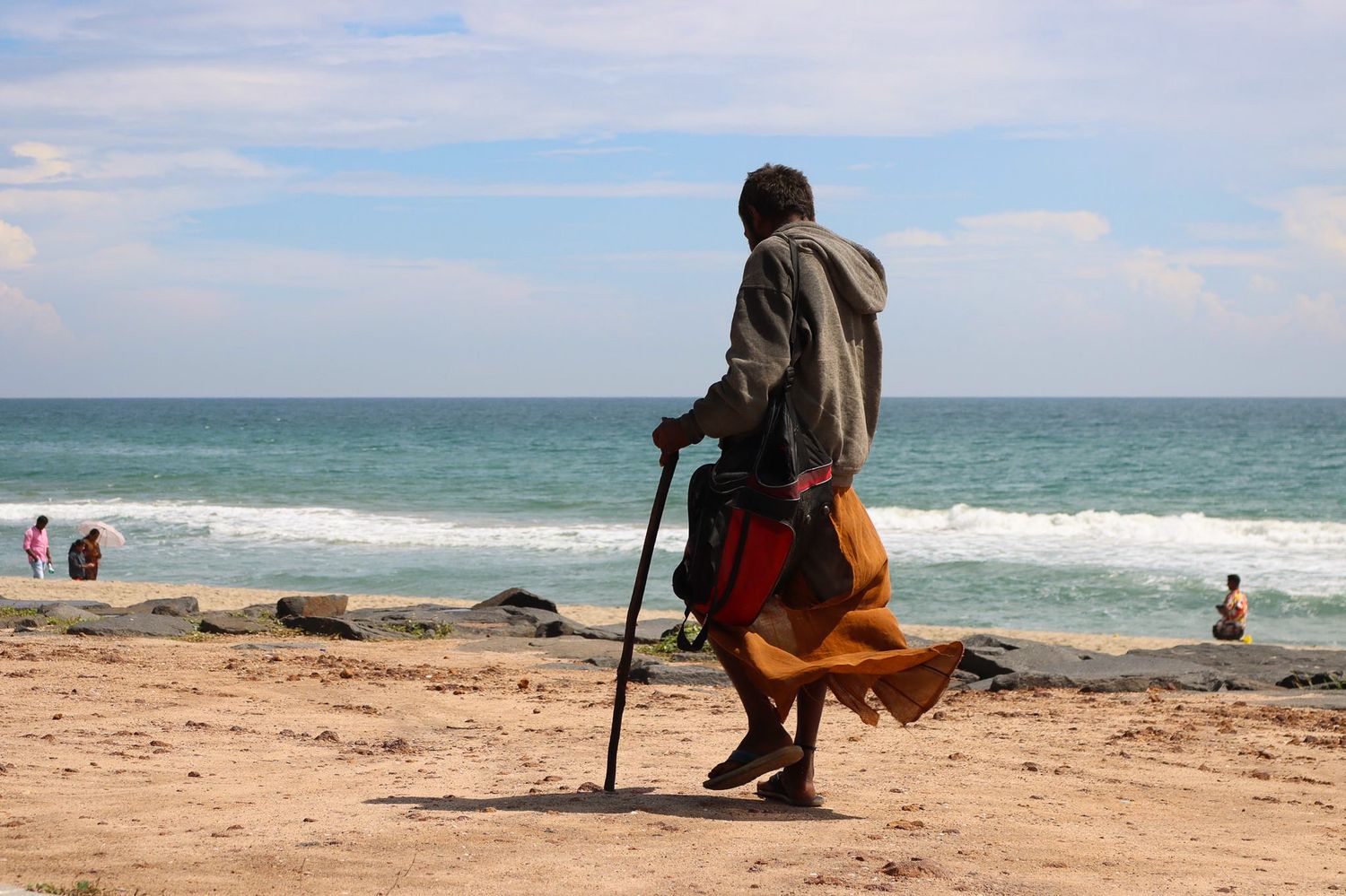 Man with a walking stick silhouetted against the sea on a sandy beach in Puducherry, India.