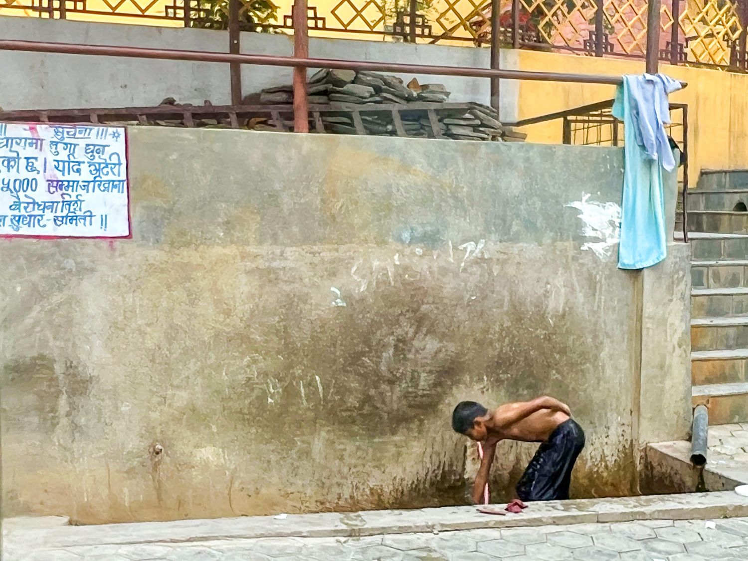 Boy bending over to wash or drink from a street-side water source next to a wall in Kathmandu, Nepal