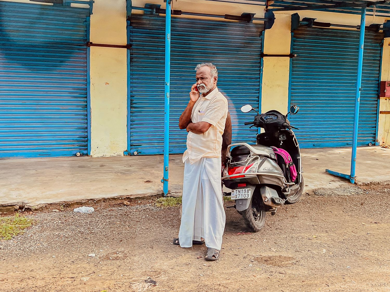 Elderly man standing beside a parked scooter on a quiet street with shuttered shops in India
