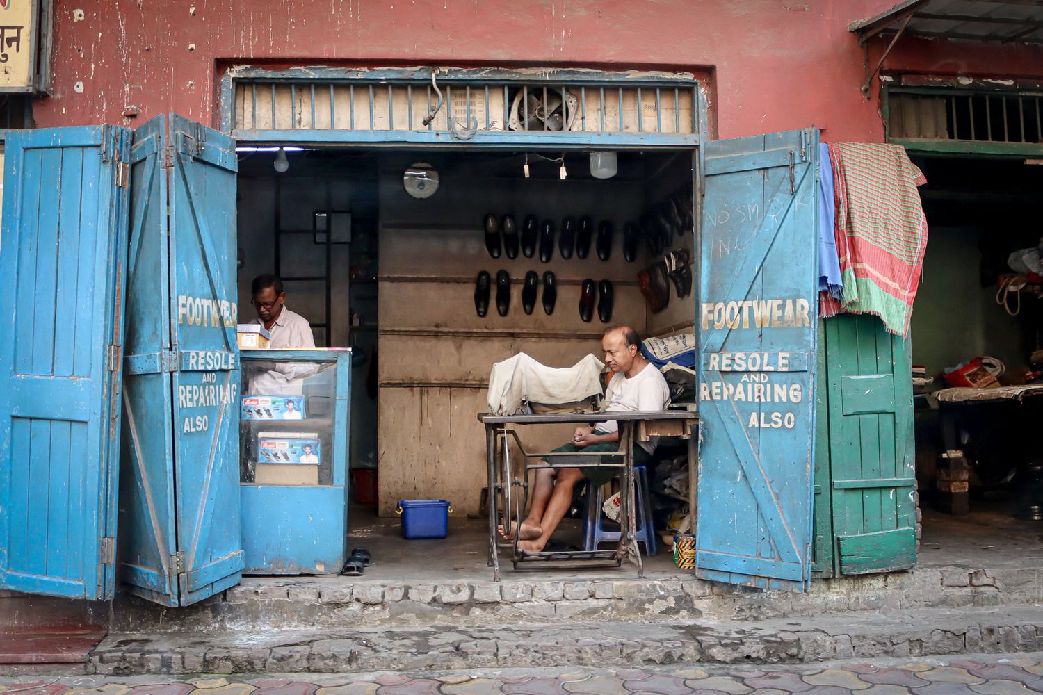 Cobbler working inside a small repair shop with blue shutters and signage. Kolkata, India