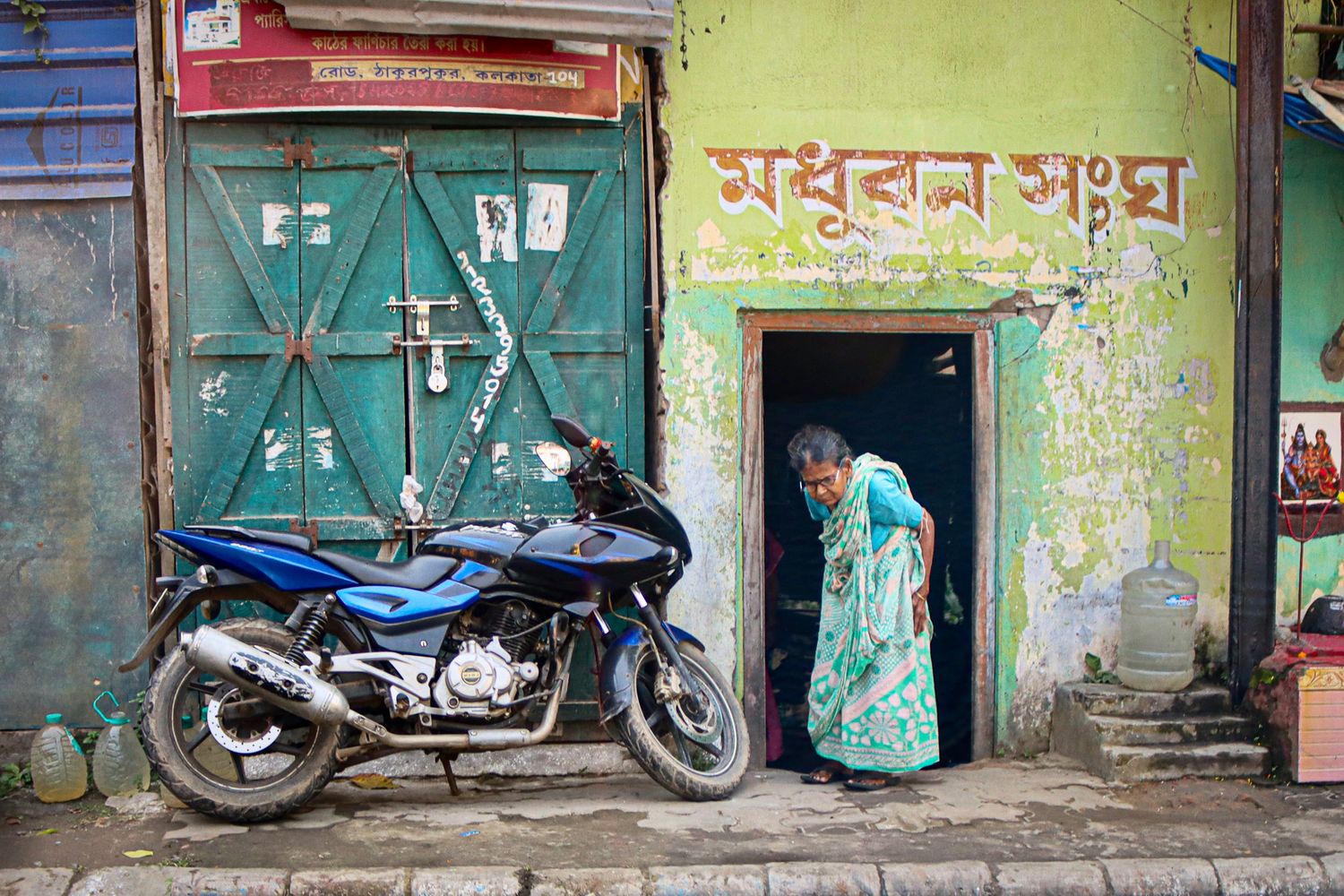 Elderly woman stepping into a doorway under weathered signage and green paint. next to a motorcycle. India