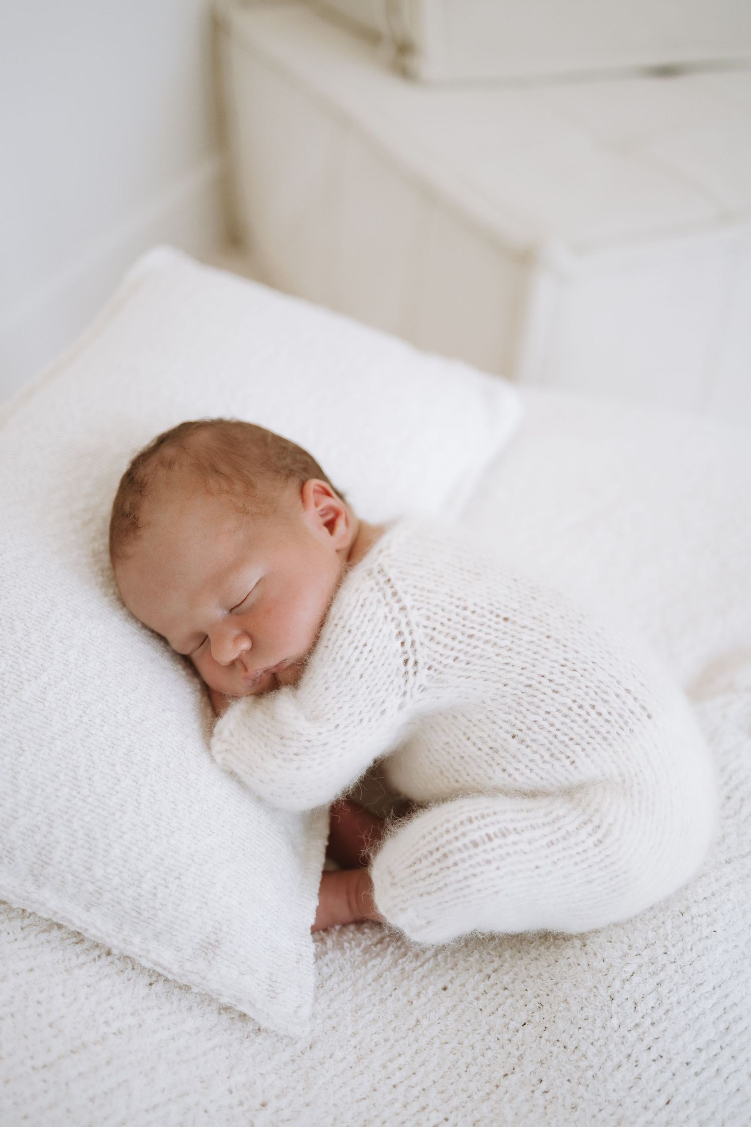 Sleeping newborn baby in white onesie rests peacefully in someone's arms.