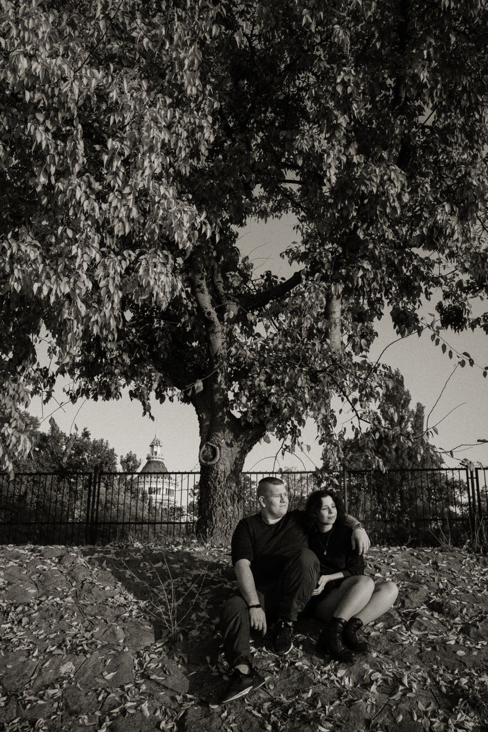 A couple sitting together under a large tree on ground covered in fallen leaves.