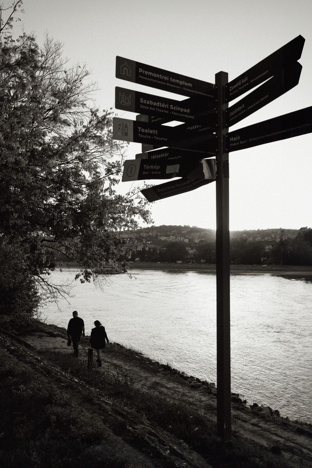 Two silhouettes walking along a riverbank path with directional signs in black and white.