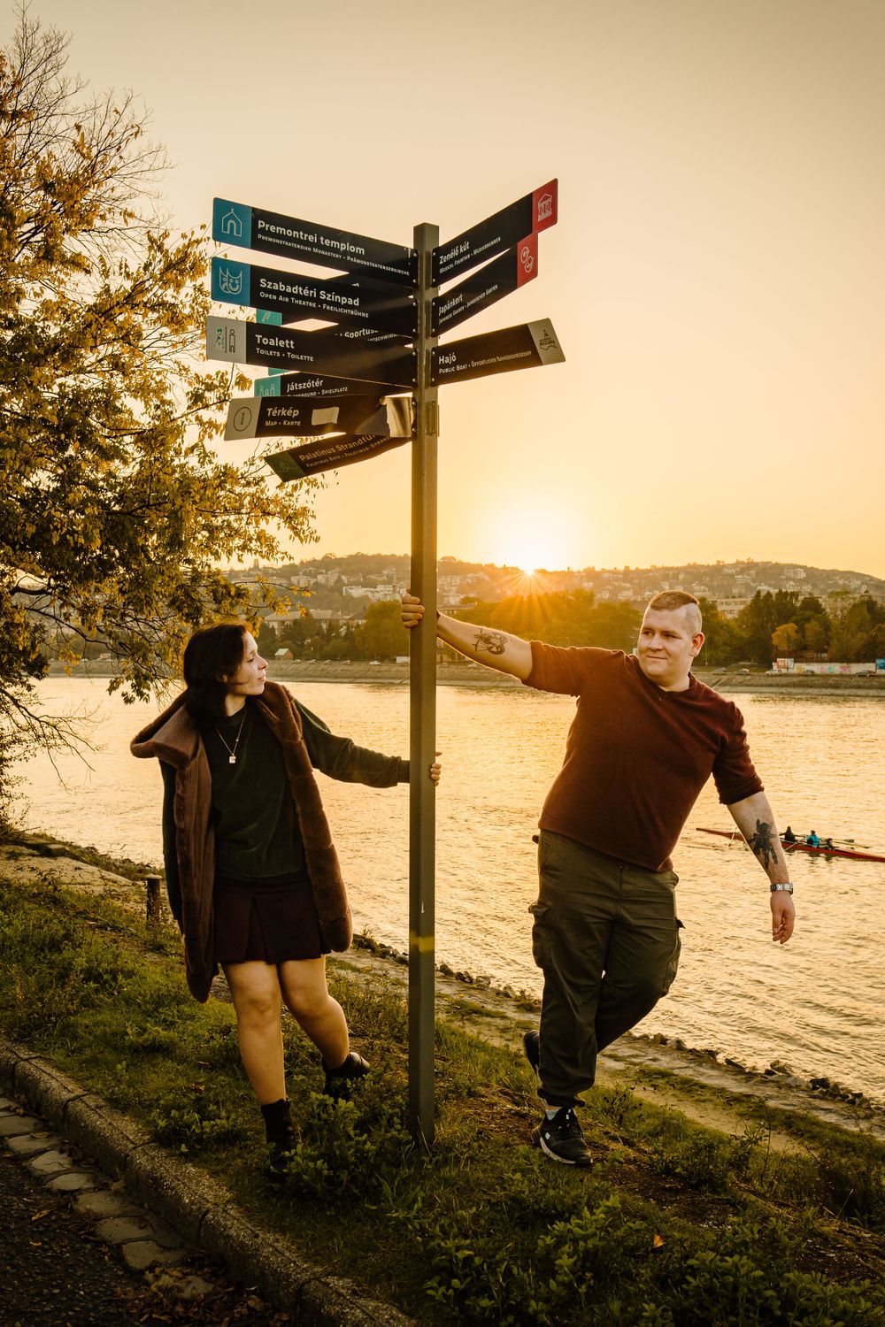 Couple posing playfully by directional sign post near waterfront at sunset.