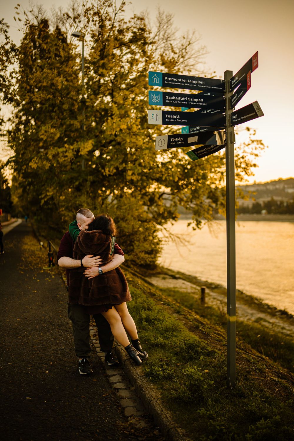 Romantic embrace by directional signpost along waterfront path at golden hour.