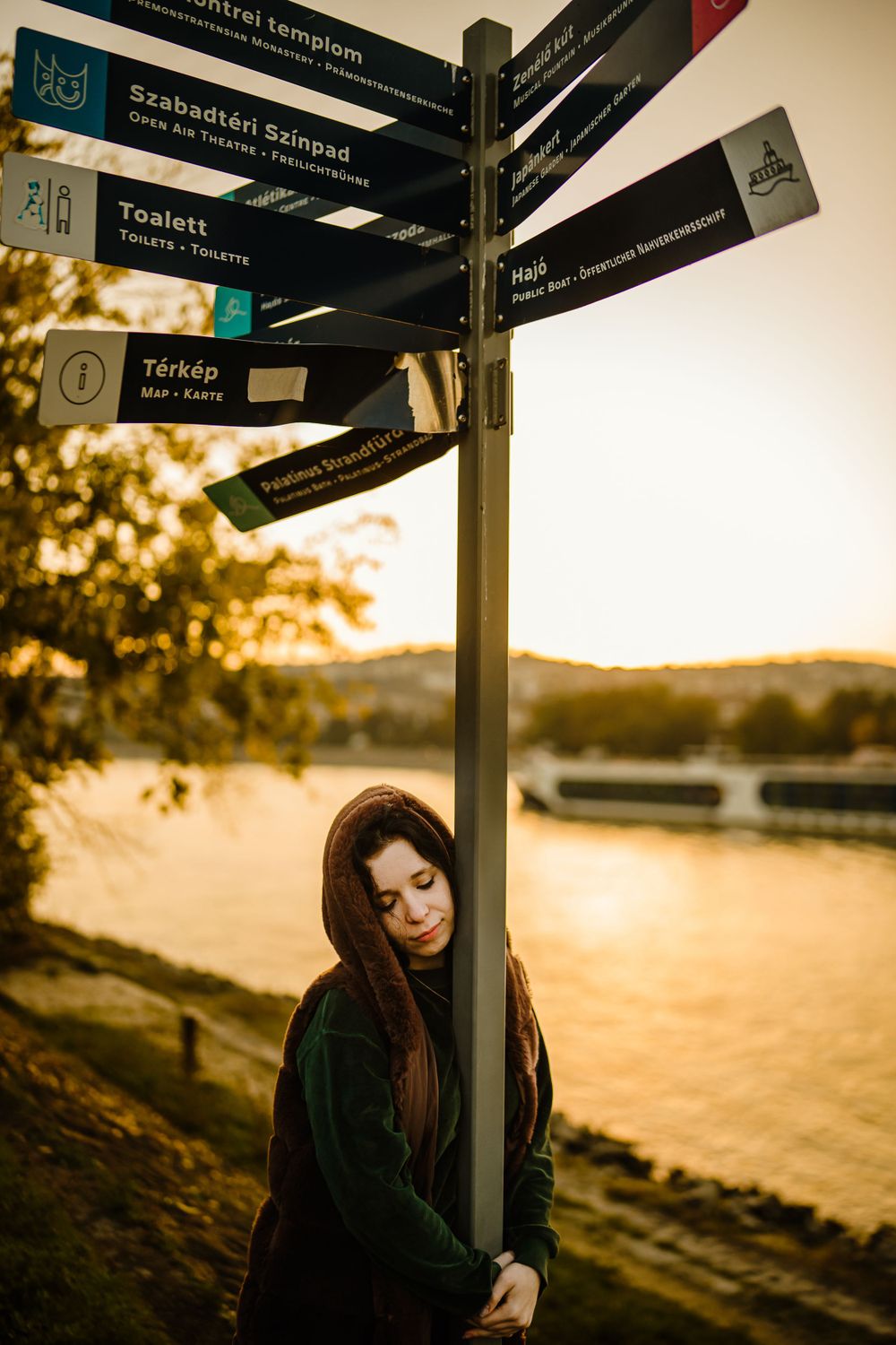 Woman in green coat leaning against directional sign post by river at sunset.