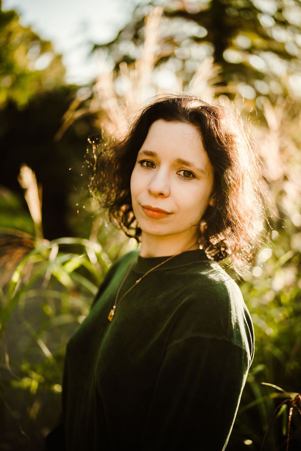 Portrait of someone in a green sweater backlit by golden sunlight among foliage.
