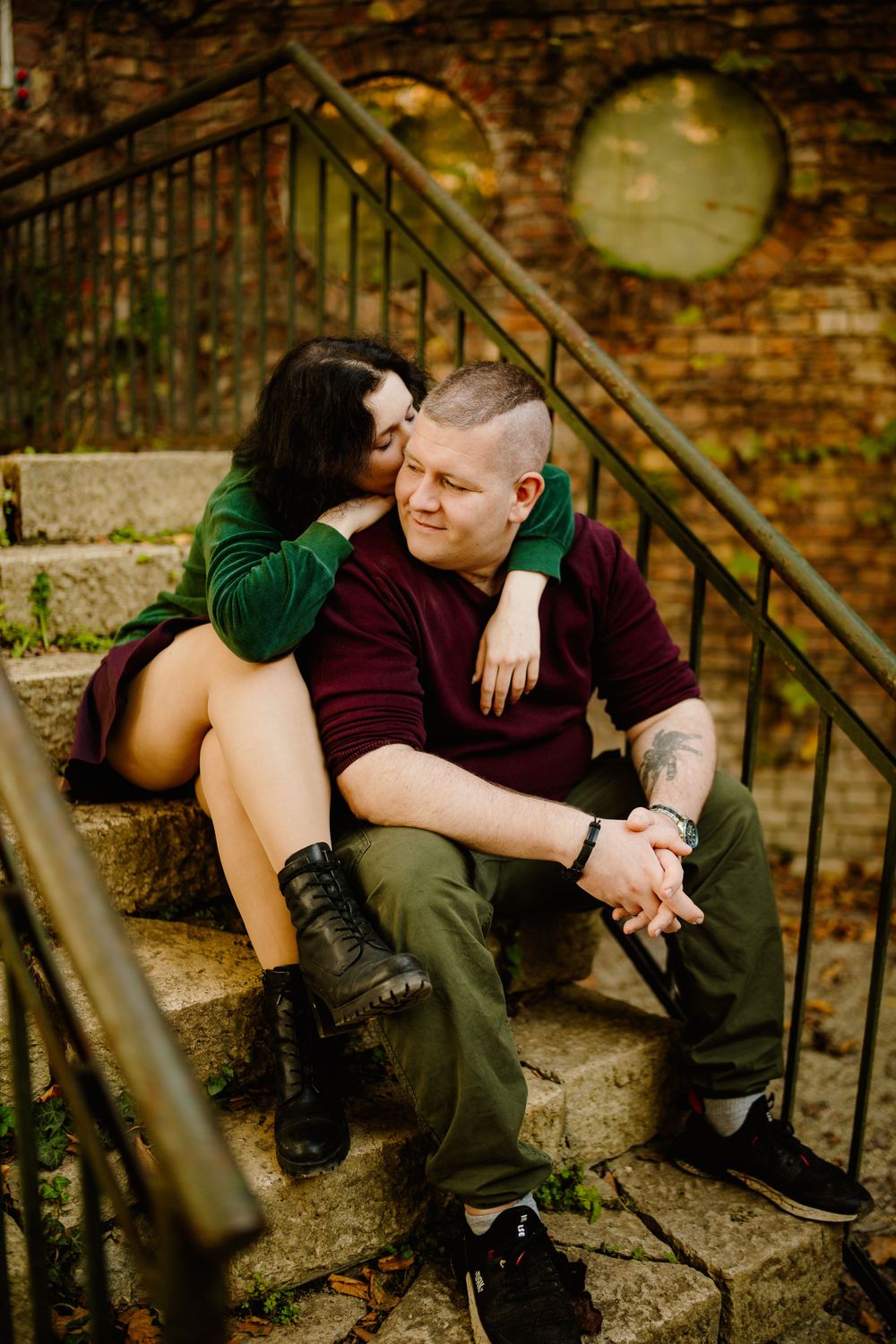 Romantic couple sitting on stone steps against rustic brick wall with circular window.