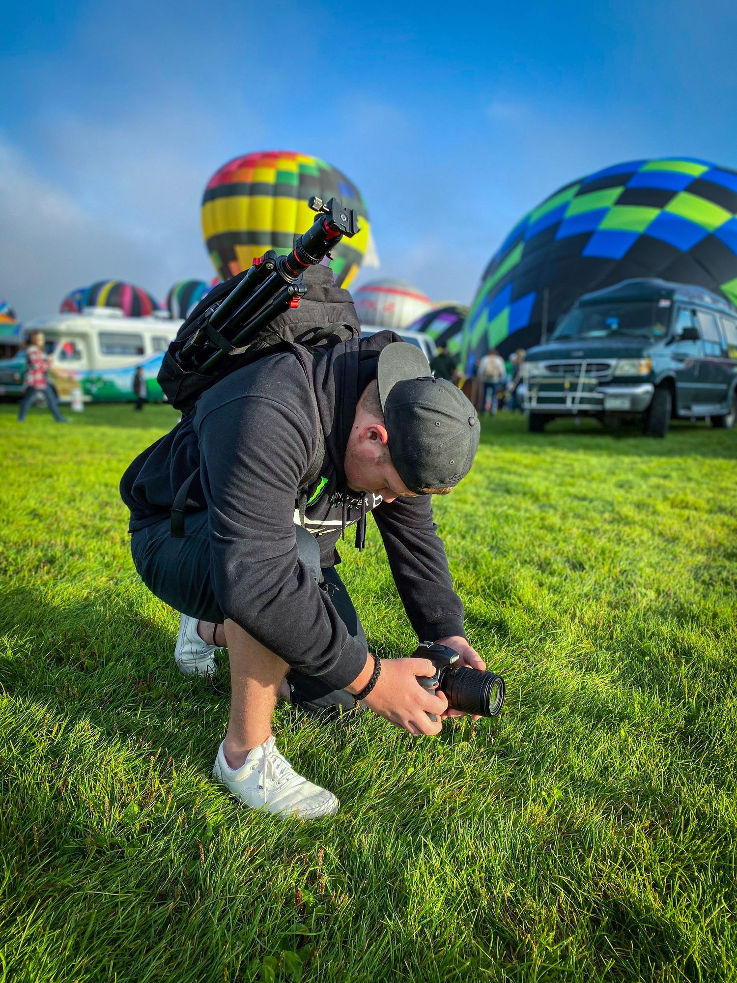 Photographer in dark jacket crouches low to capture hot air balloons at an outdoor event on grassy field.