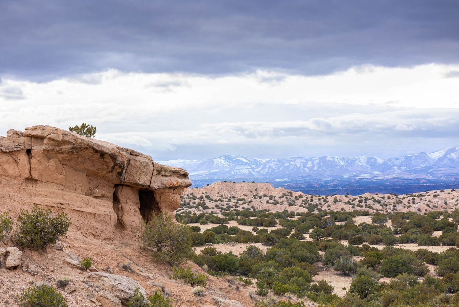 Desert landscape with red rock formations and snow-capped mountains under dramatic cloudy skies.