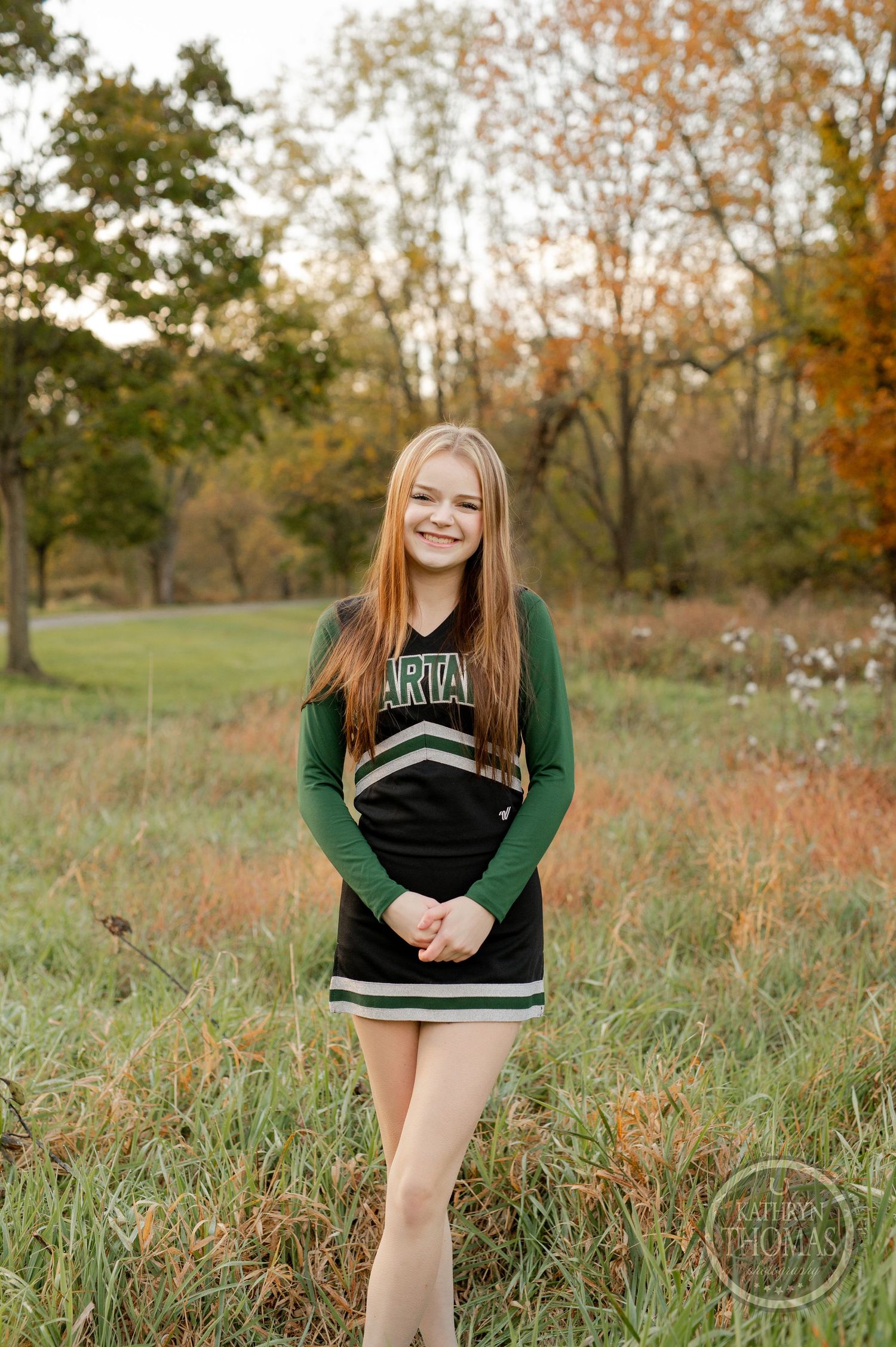 Cheerleader stands in tall grass field wearing green and black uniform during golden hour.