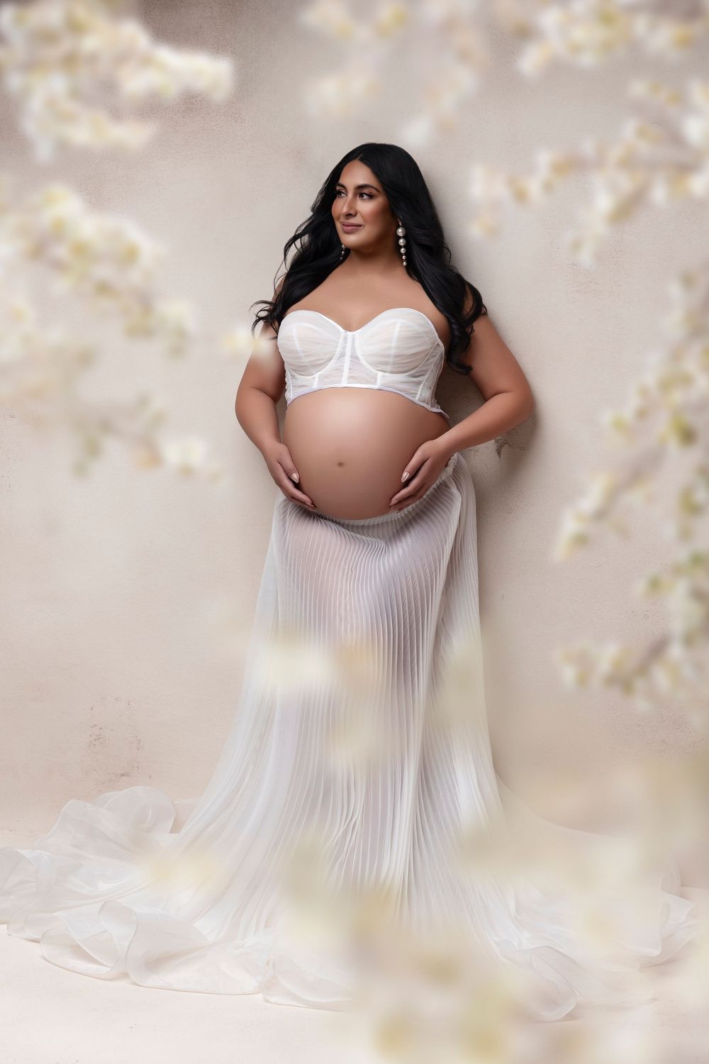 Pregnant woman in white bustier and sheer pleated skirt cradling baby bump, surrounded by soft floral backdrop.