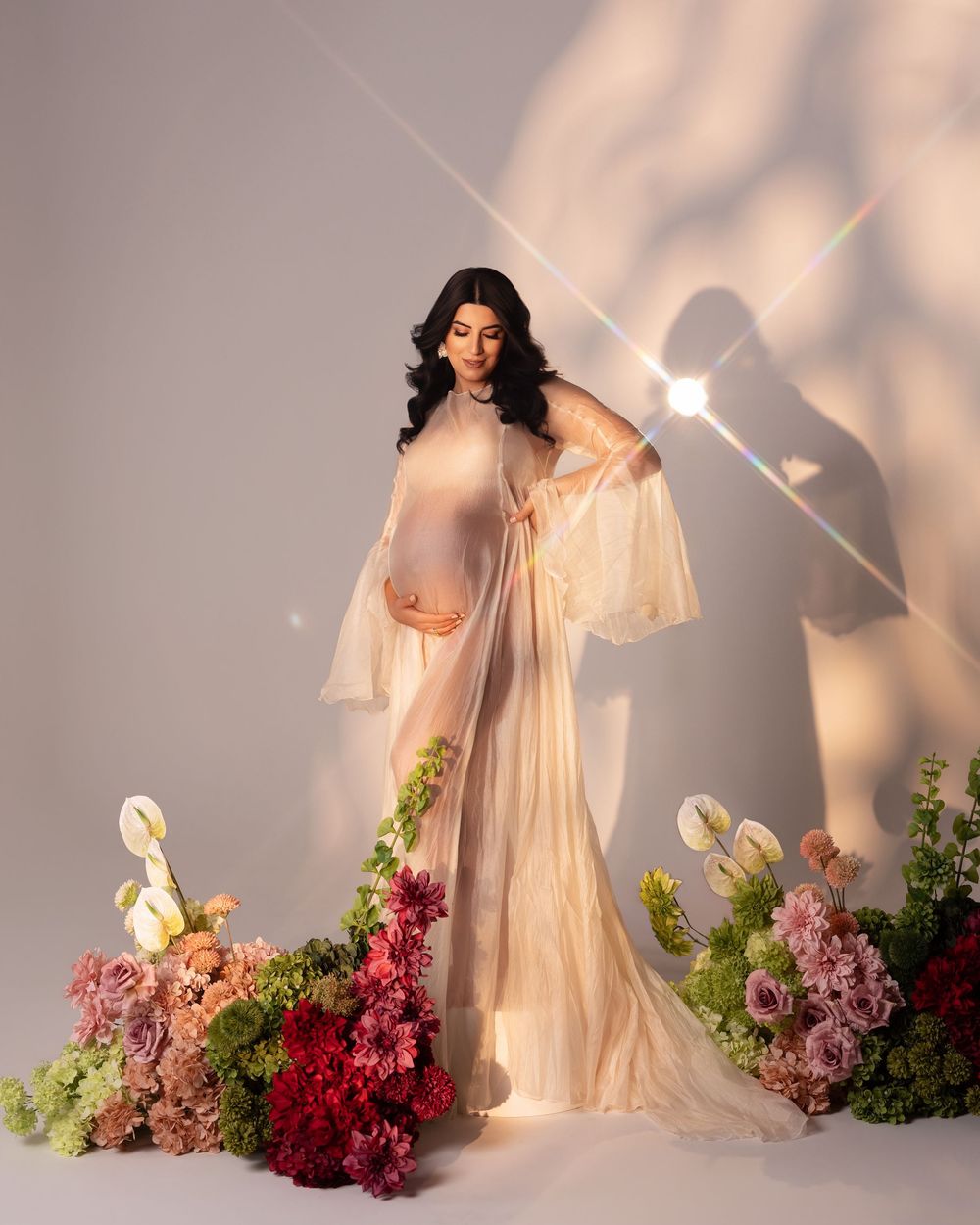 Pregnant woman in sheer cream gown surrounded by colorful flowers in an elegant maternity photography studio shoot.