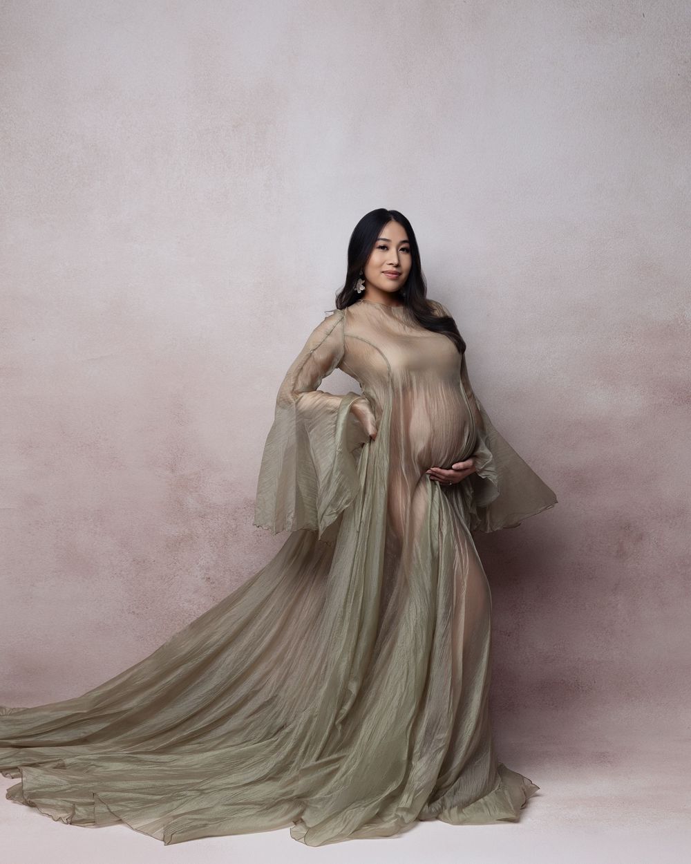 Pregnant woman in flowing sage green sheer maternity gown posing gracefully in a professional studio setting.