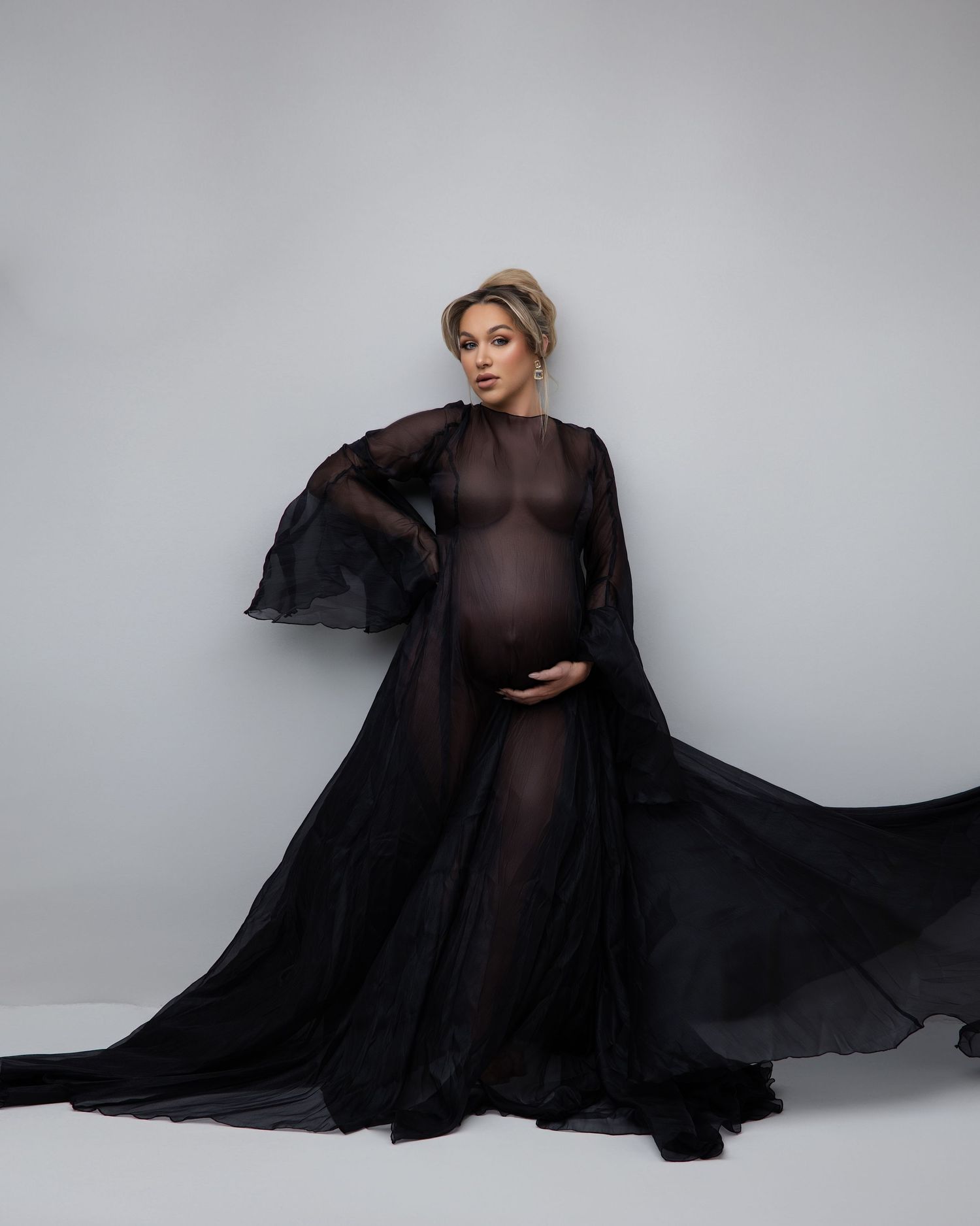 An elegant maternity portrait with a flowing black sheer gown and cape against a light gray studio backdrop.