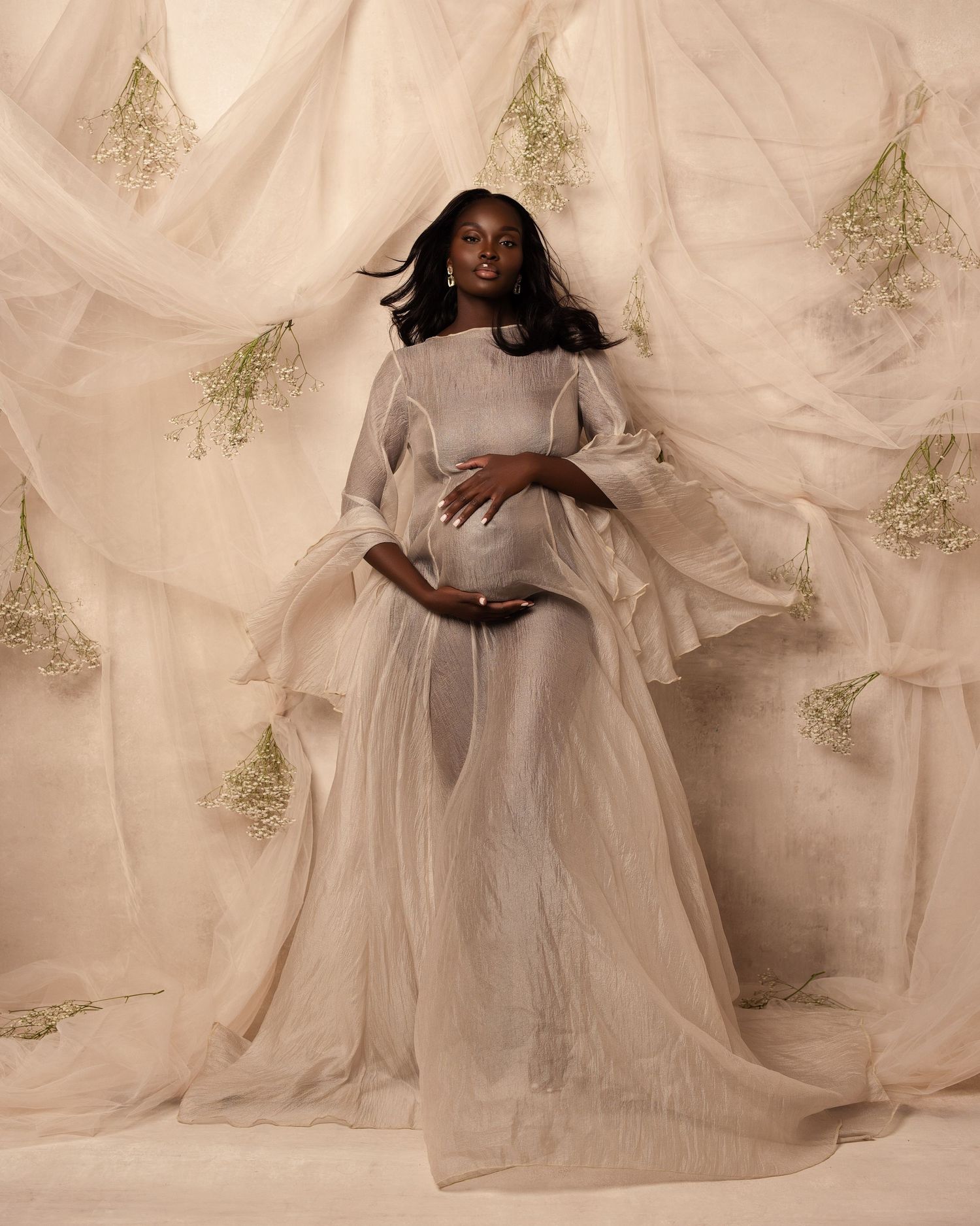 Ethereal maternity photo with flowing white gown and sheer fabric backdrop adorned with delicate gold foliage details.