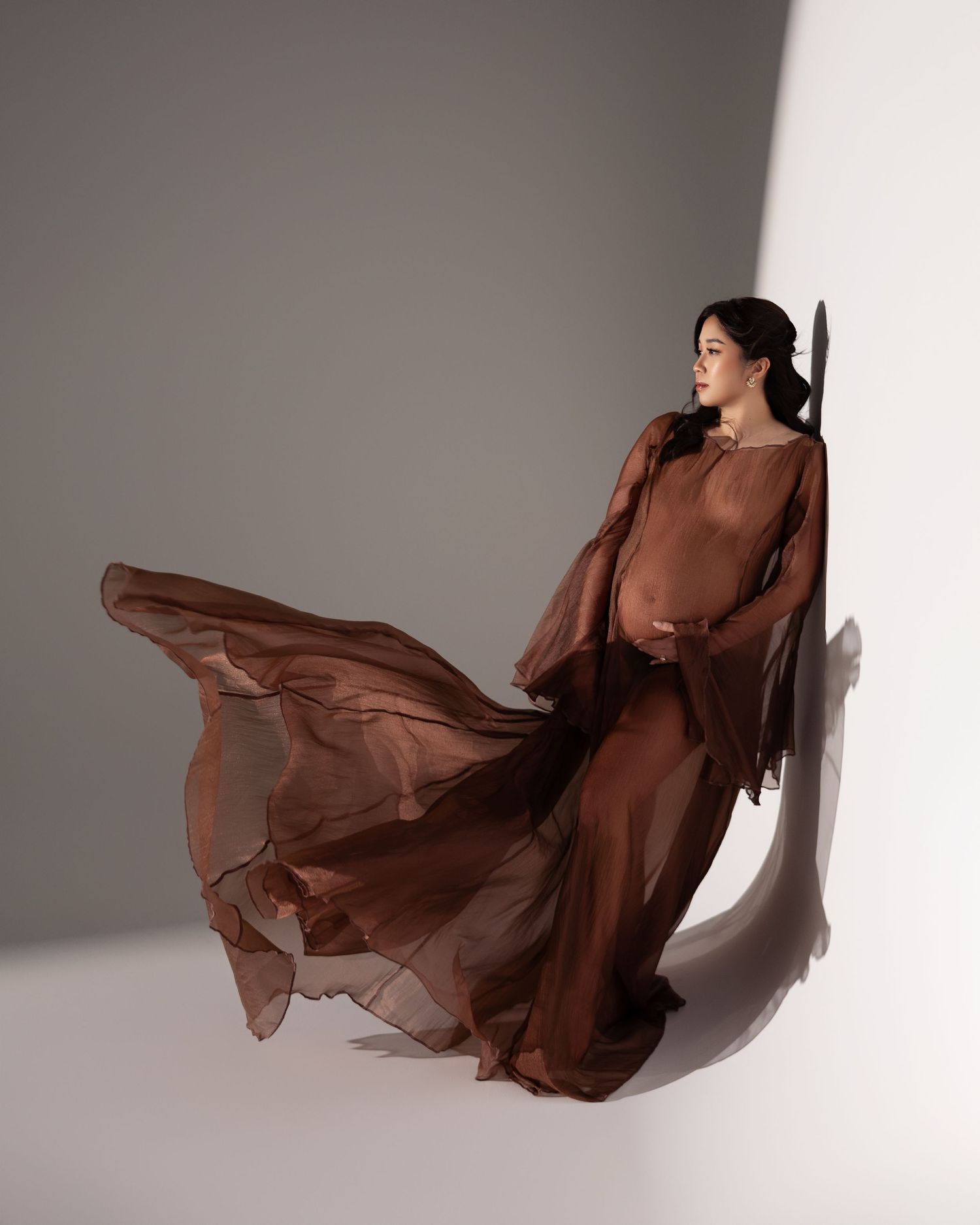 Flowing brown chiffon maternity dress with dramatic movement captured in a minimalist studio setting with shadows.