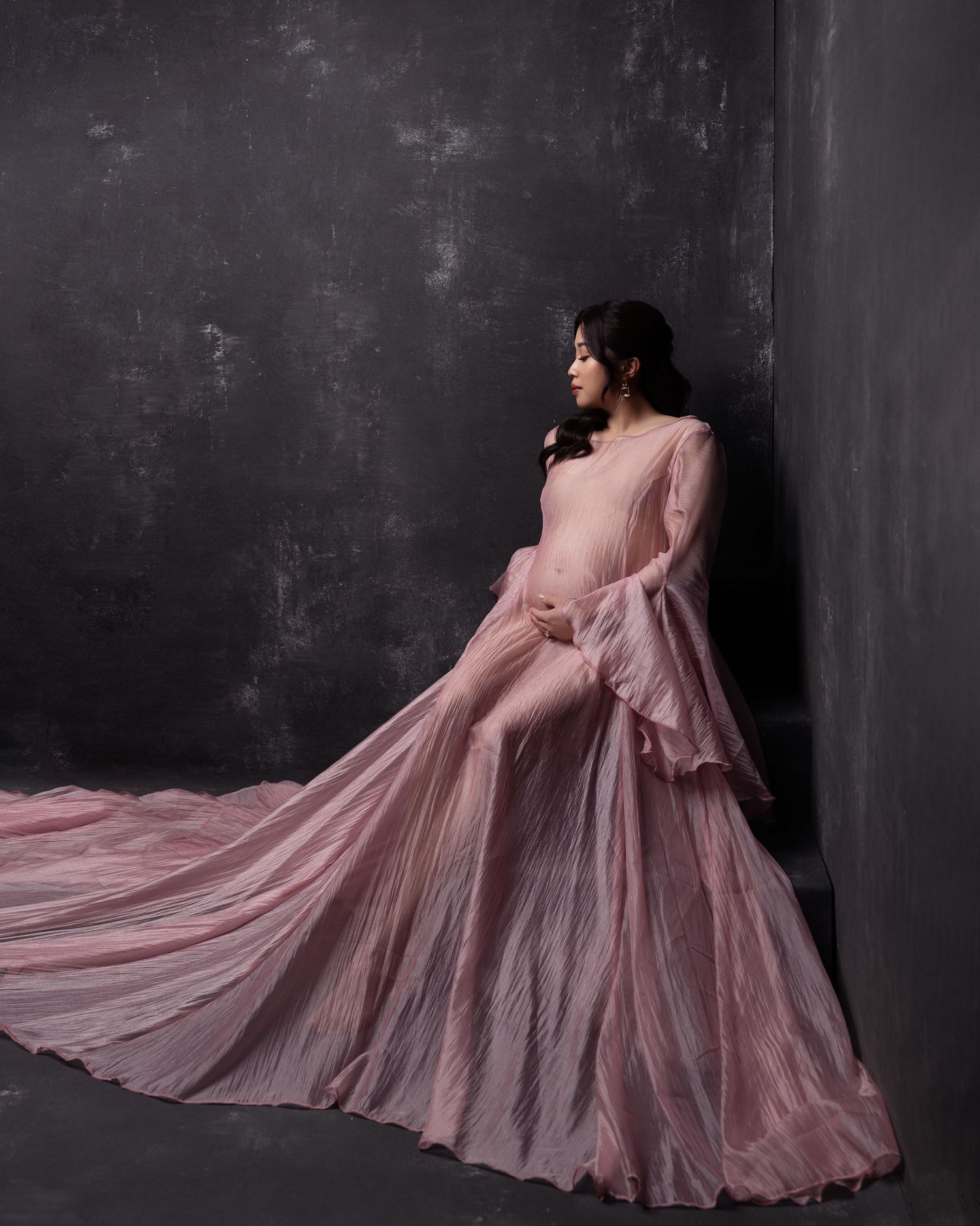A person in a flowing dusty pink gown stands against a dark wall, with the dress fabric dramatically billowing outward.
