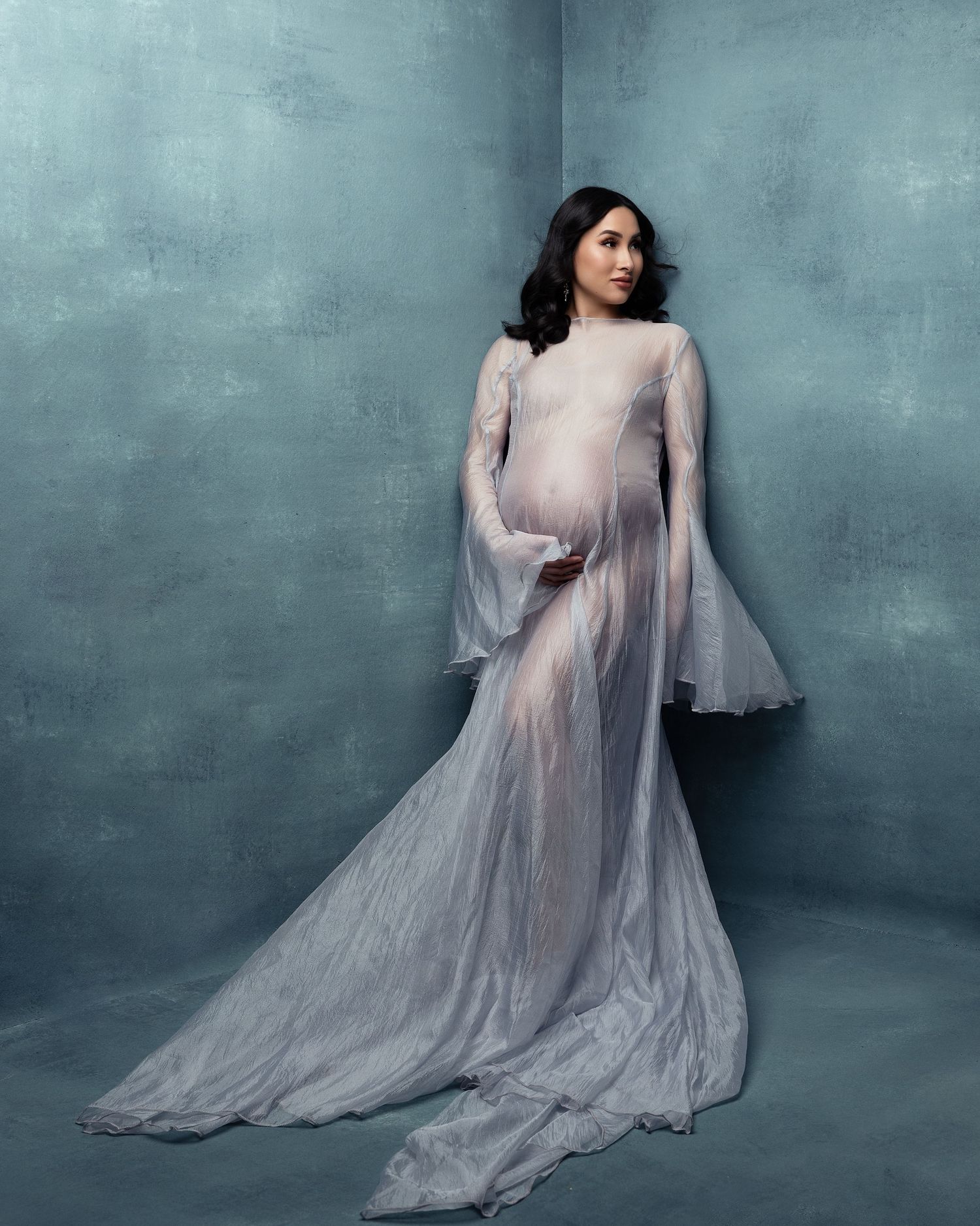 A woman in an ethereal sheer white maternity gown with bell sleeves poses against a teal blue textured backdrop.