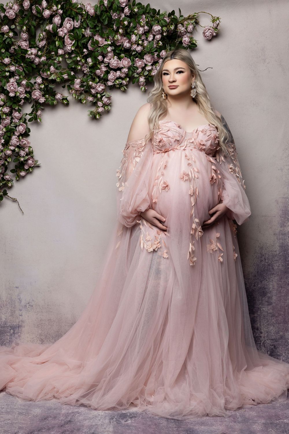 Ethereal maternity gown in blush pink tulle with floral appliques and flowing sleeves against climbing roses backdrop.