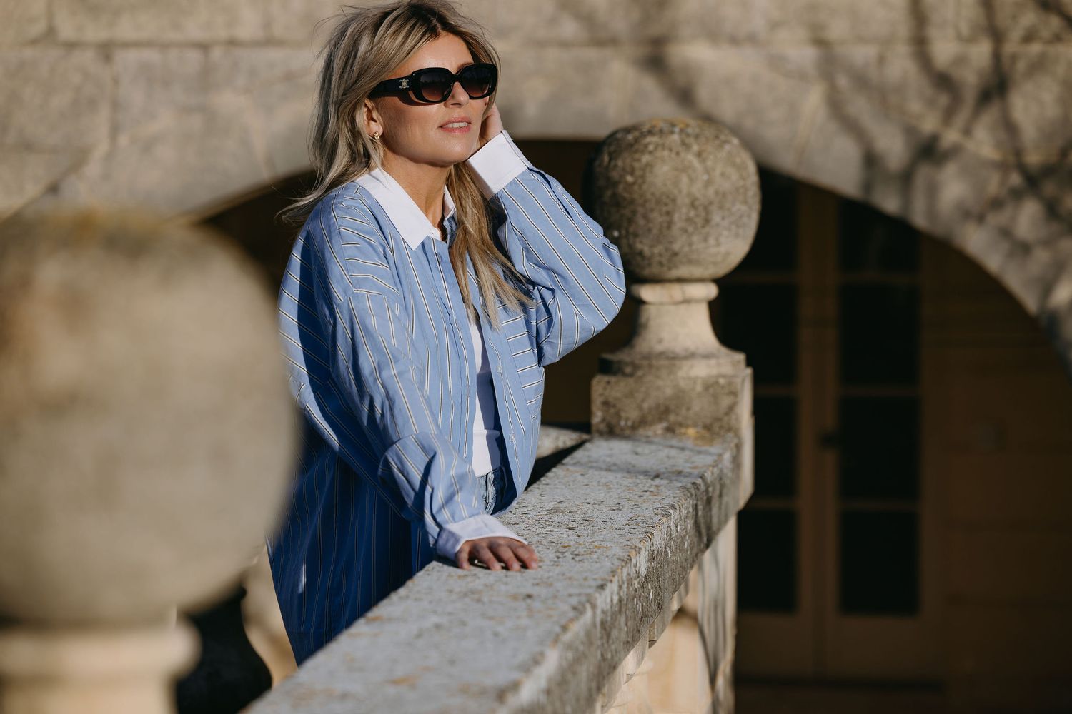 Woman in denim shirt leaning on stone balcony in sunlight.