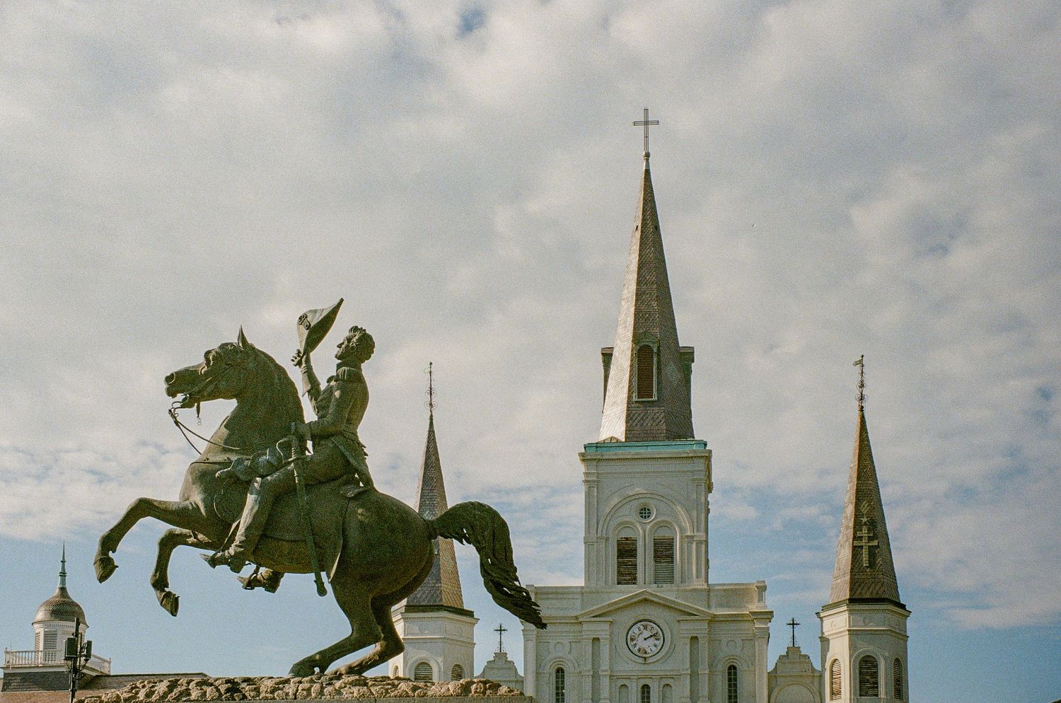 An equestrian statue stands dramatically in front of a white church with twin spires against a cloudy sky.